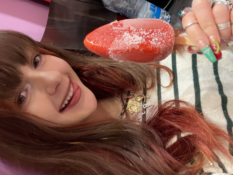 A smiling woman with brown hair and bangs, some red streaks, and tinsel strands, holds a frozen red ice pop with a wooden stick. The woman has brown eyes and shows her teeth with her tongue slightly visible. She has multiple rings on her fingers with colorful acrylic nails. She wears a necklace with multiple charms including a Star of David. A plastic water bottle is in the background. The background has purple and white-green stripes.