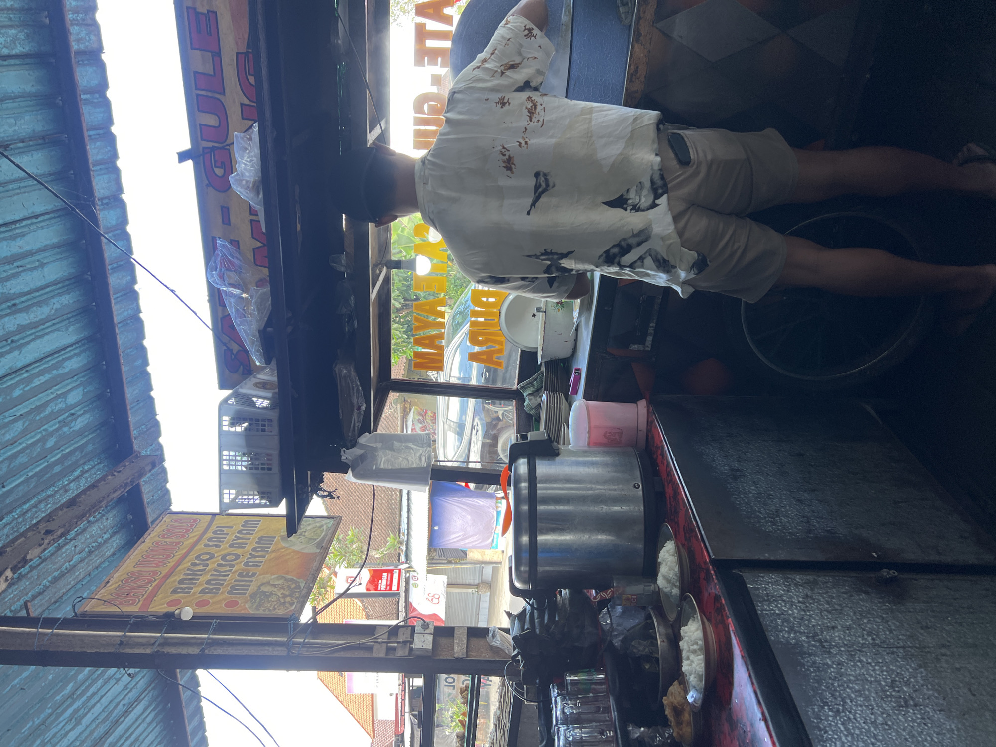 The image depicts an outdoor food stall in Payangan, Indonesia, likely a local eatery. A man wearing a patterned short-sleeved shirt and beige shorts stands with his back to the camera, appearing to be preparing food. He is positioned in front of a stall counter laden with various items, including a large metal rice cooker, plates of rice, glasses, and what seem to be food ingredients in containers.
Behind the man, a window reveals the street scene of Payangan. There are buildings with tiled roofs, some with promotional banners, and parked vehicles. One banner prominently displays "BAKSO WONG SOLO" and lists menu items like "BAKSO SAPI," "BAKSO AYAM," and "MIE AYAM," indicating that this stall specializes in meatballs and noodle dishes. Another banner visible in the background reads "SATE GULE," suggesting satay and gulai are also offered.
The stall itself is covered by a corrugated metal roof, providing shelter from the elements. The overall atmosphere suggests a busy, functional food service environment common in a tropical climate. The lighting indicates it is daytime, with the sun casting shadows and highlighting the textures of the stall and its surroundings.