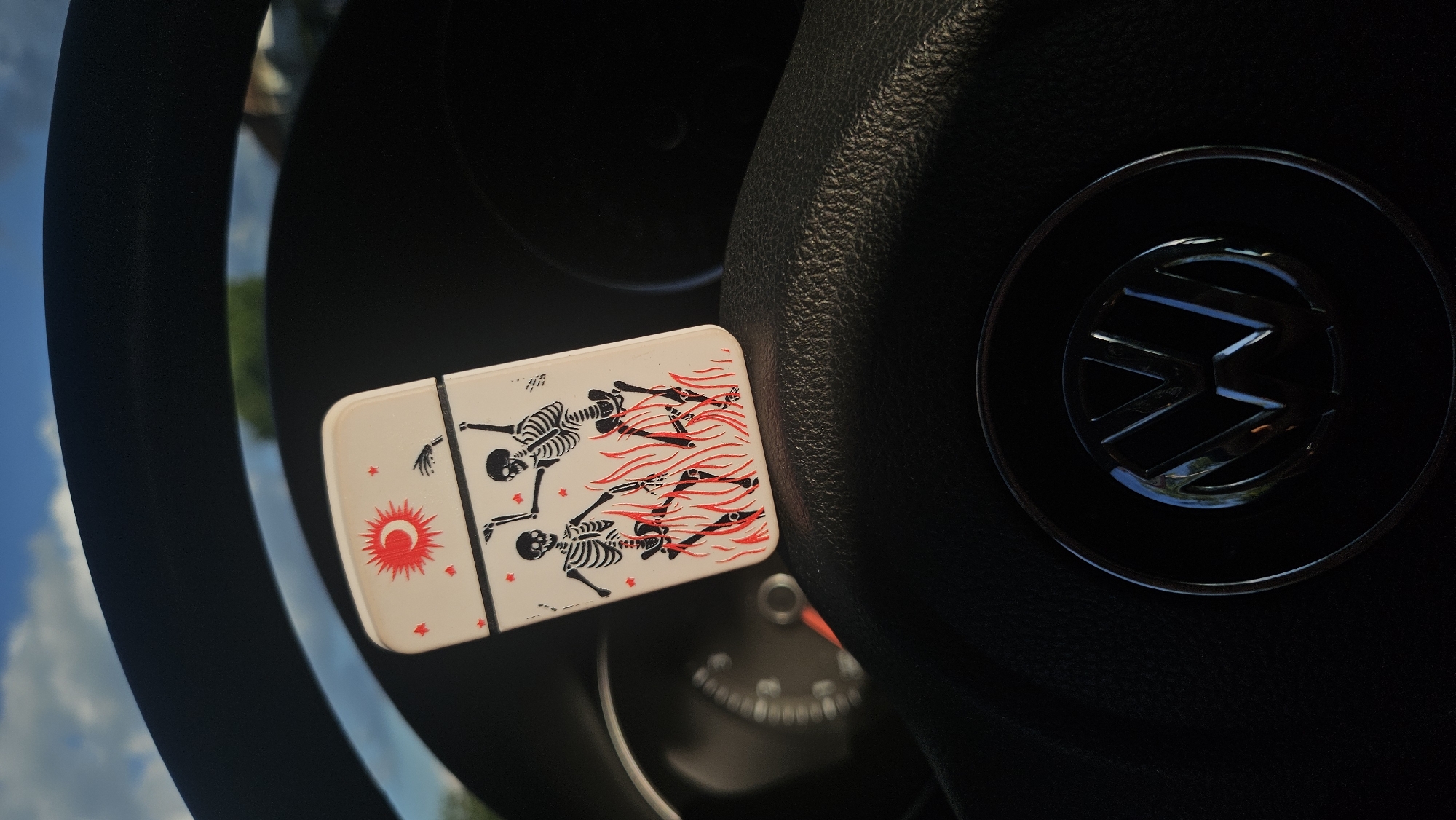 This image shows a close-up view of a steering wheel in a car, with a decorative item placed on it. The steering wheel is black and appears to be made of leather. In the center of the steering wheel is the Volkswagen logo, a chrome emblem with the letters "VW" intertwined within a circle.
On the steering wheel, to the left of the logo, is a rectangular, white object with a decorative design. The design features several black skeletons in various poses, some appearing to be dancing or running. They are depicted with detailed skeletal structures. Below the skeletons on the left side of the object, there is a red circular symbol, possibly representing the sun or a stylized moon, surrounded by small red stars. The rest of the object's surface has wavy red lines, suggesting movement or flames.
The background of the image is blurred but suggests an outdoor scene. Parts of a blue sky with white clouds and some greenery are visible around the edges of the steering wheel. The lighting indicates it is daytime, likely sunny given the bright sky. The overall impression is a casual snapshot of a car interior with a unique decorative element. There is no visible text in the image other than the "VW" logo.