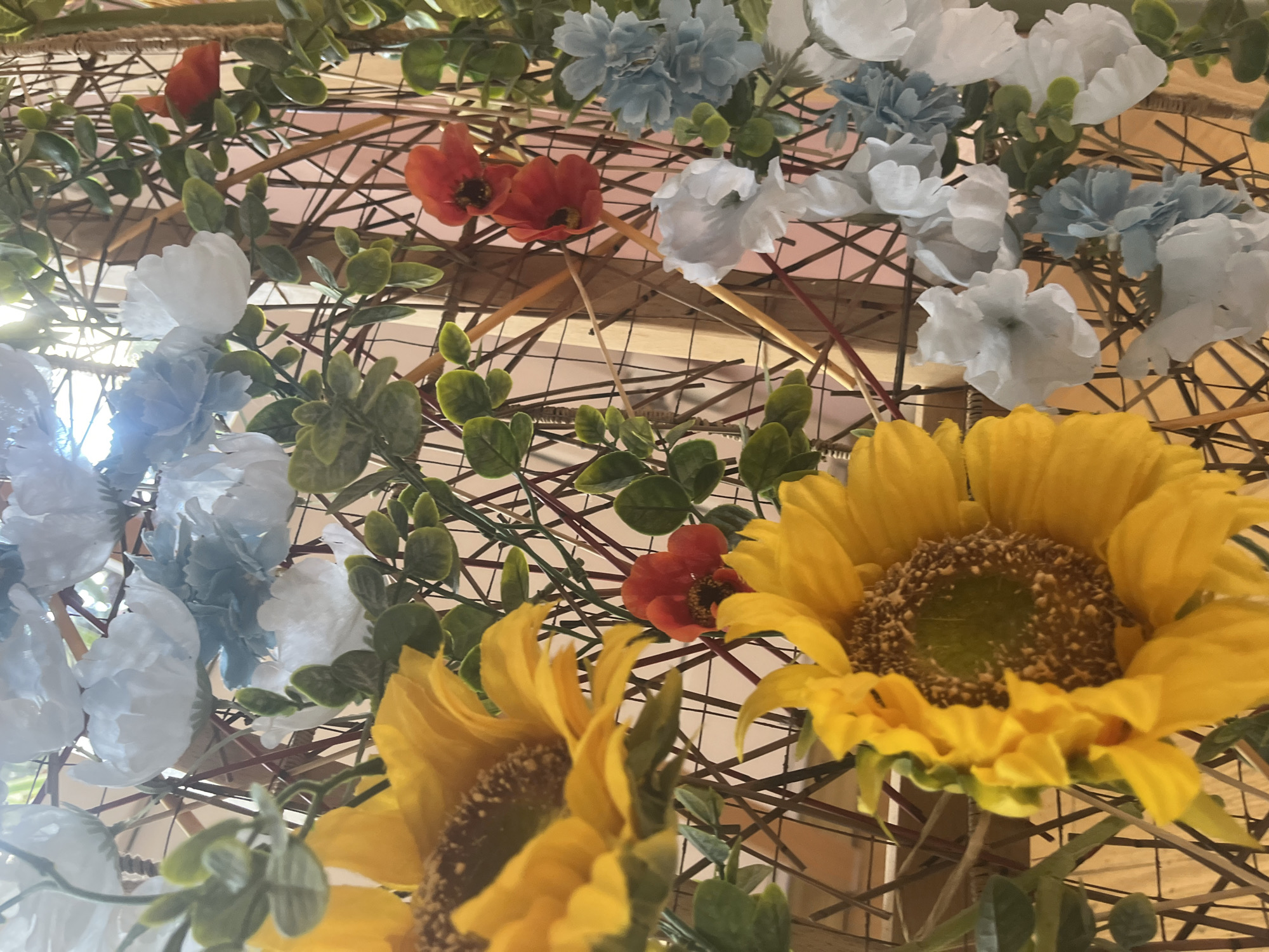 The image depicts a floral arrangement against a backdrop of woven wooden or bamboo slats. Two sunflowers, one partially obscured, dominate the lower portion of the frame. Above, there are interspersed artificial flowers in shades of white, light blue, and a few red or orange blossoms. Green foliage is mixed among the flowers and structural lattice. The background is nondescript and offers no clues to suggest a specific location, including the city of Saint Petersburg, Russia. The artificial nature of the flowers suggests this is a decorative display, likely indoors.