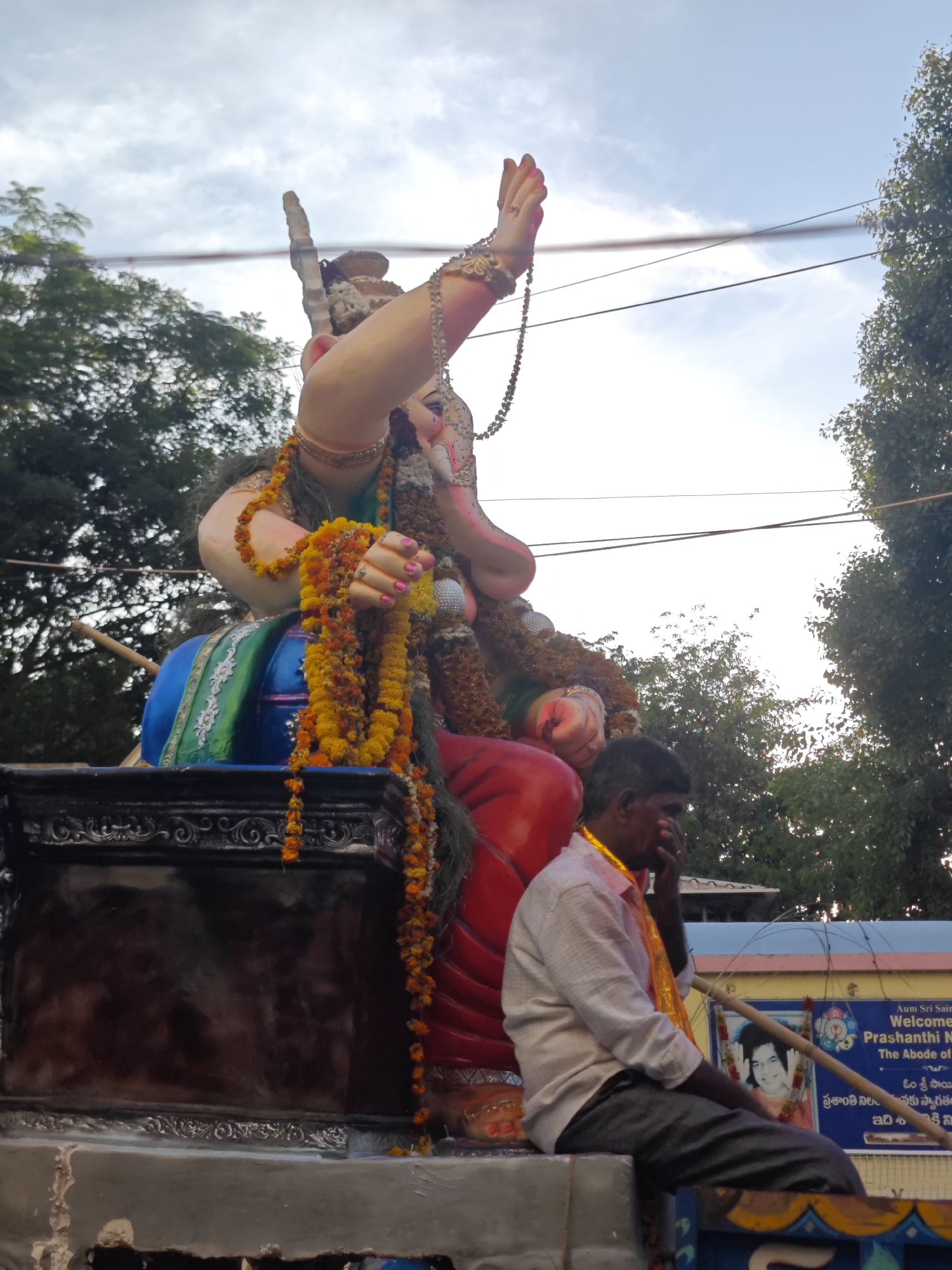 A full shot captures a large statue of the Hindu deity Ganesha, adorned with orange and yellow marigold garlands, being paraded on a vehicle. The statue is brightly colored with a red lower body and a blue and green upper body, decorated with ornate jewelry and necklaces. In the foreground, a man with dark skin and short black hair sits on the edge of the vehicle, wearing a light-colored, long-sleeved shirt and dark pants. He is touching his face, appearing to be wiping his eyes or nose. Behind him, a banner displays text in English and Telugu, welcoming visitors to "Prashanthi Nilayam, The Abode of Peace" in Puttaparthi, India. The background features lush green trees and a cloudy sky. The scene appears to be a religious procession or festival.