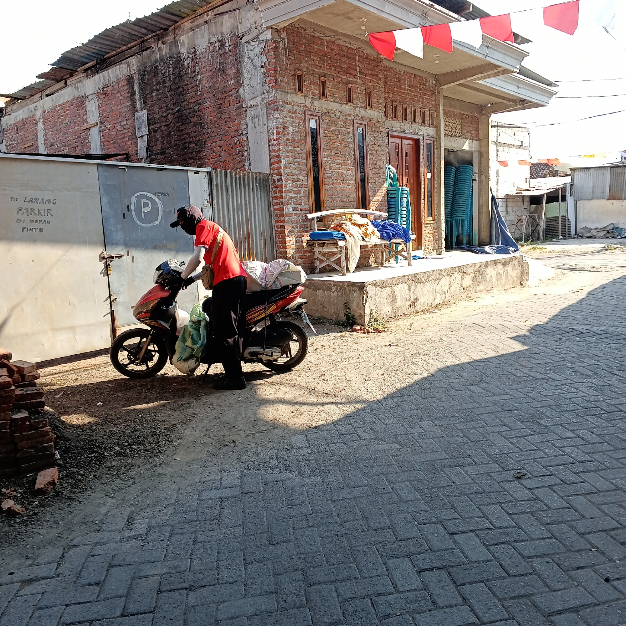 In Kepuh Kiriman, Indonesia, a person wearing a red shirt, black pants, and a black cap with a face covering stands next to a red and black motorcycle. The motorcycle has a white and green bag on its rear seat. The person appears to be interacting with the motorcycle, possibly preparing to depart. Behind them, a grey metal gate is visible with writing that translates to "DO NOT PARK IN FRONT OF THE DOOR" and a parking symbol. To the right of the gate, a brick building with a white overhang is present, adorned with red and white Indonesian flags. The ground is paved with grey bricks. In the background, more buildings and a blue tarp are visible. The scene is illuminated by sunlight, casting shadows on the ground.