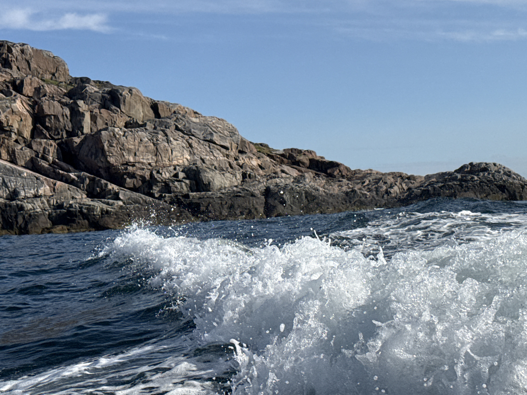 This is a medium shot of a rocky coastline with waves crashing. The rocks are dark and jagged, with some patches of green vegetation. The water is a deep blue, and the waves are white and foamy. The sky is clear and blue with a few wispy clouds. The image is taken from a low angle, as if from a boat. There are no people or man-made objects visible in the image. The location is not specified and cannot be determined from the image.