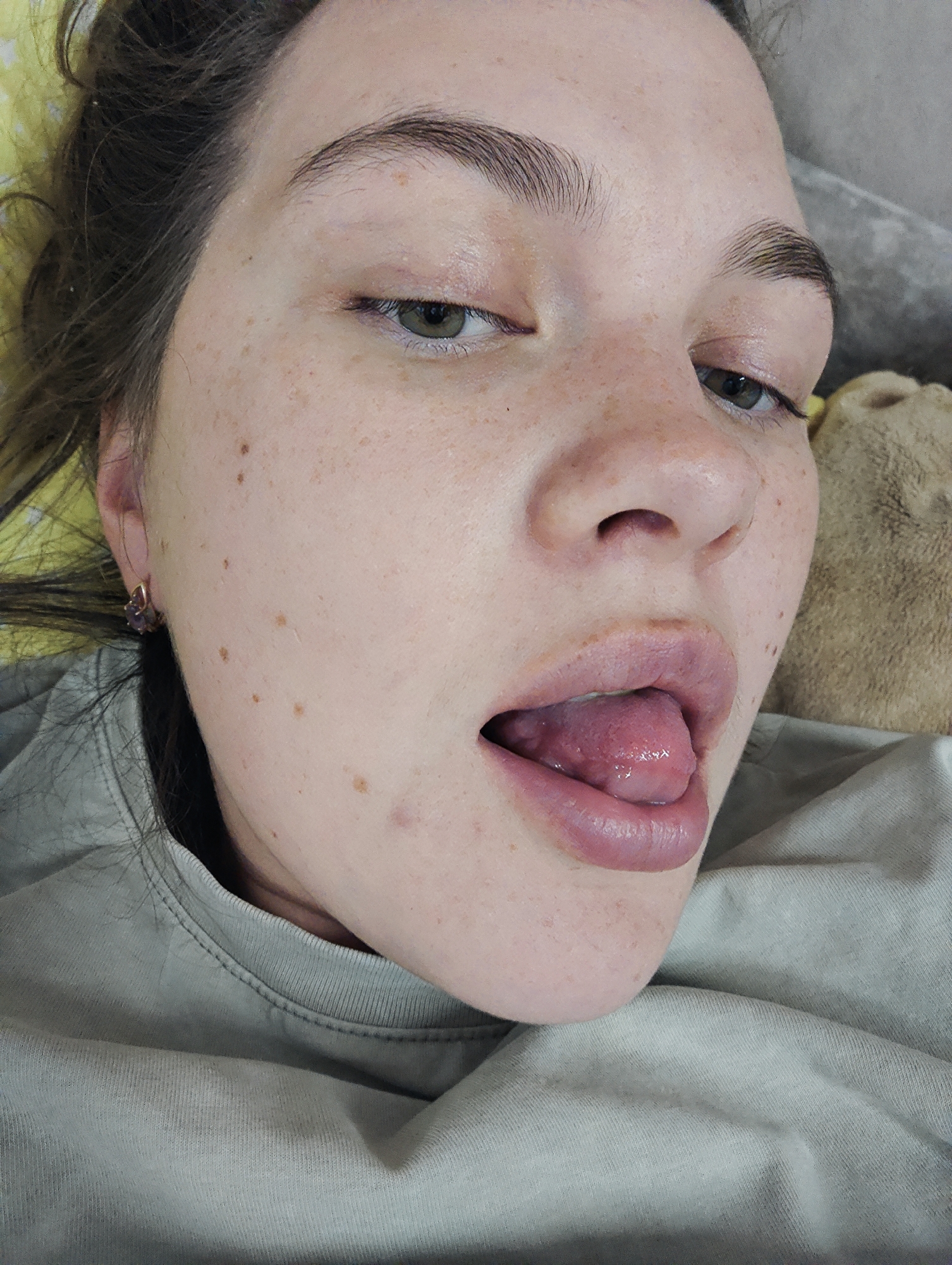 A close-up, slightly angled portrait captures a young woman lying down in an indoor setting in Chaikovsky, Russia. Her face, positioned prominently in the foreground, is fair-skinned and adorned with numerous freckles and a few moles. She has dark, full eyebrows, and her greenish-brown eyes are partially closed or looking downward. Dark brown hair frames her face and is visible on the left side of the frame, appearing somewhat dishevelled.
Her full, pinkish lips are slightly parted, revealing her tongue partially protruding, which suggests a relaxed or playful expression. A small, multi-faceted purple stone earring with a gold-colored setting is visible on her left earlobe. She is wearing a light grey-green t-shirt with a ribbed crew neck collar. In the background, a hint of a yellow surface, possibly a pillow, is visible behind her head, and a soft, light brown, furry texture can be seen on the right side of the frame, indicating a comfortable indoor environment. The lighting is soft and even, highlighting her features. No visible text is present in the image.