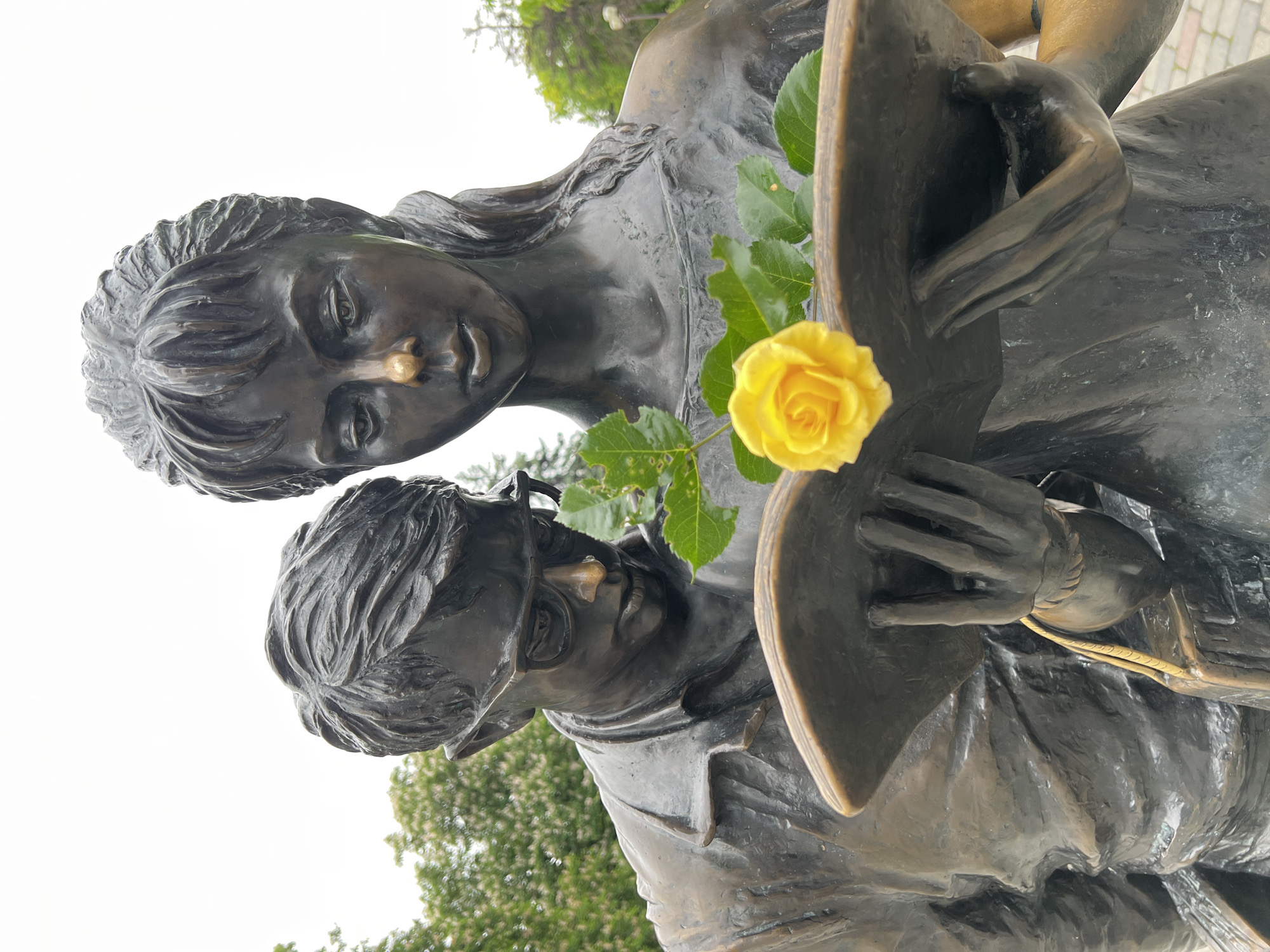 A close-up, low-angle shot depicts a bronze statue of two figures. The statue appears to be outdoors, with trees and foliage visible in the background.
The upper figure is a young woman with long hair, wearing what seems to be a simple dress. Her gaze is directed downwards. The lower figure is an older man with short hair and glasses, also looking downward. Both figures appear to be engrossed in some activity or object. They are each adorned with a small piece of gold color metal on the tip of their noses.
Between the figures, a yellow rose is positioned, adding a touch of color to the bronze scene.
The statue is the "Dog's Capital" sculpture in Krasnodar, Russia.