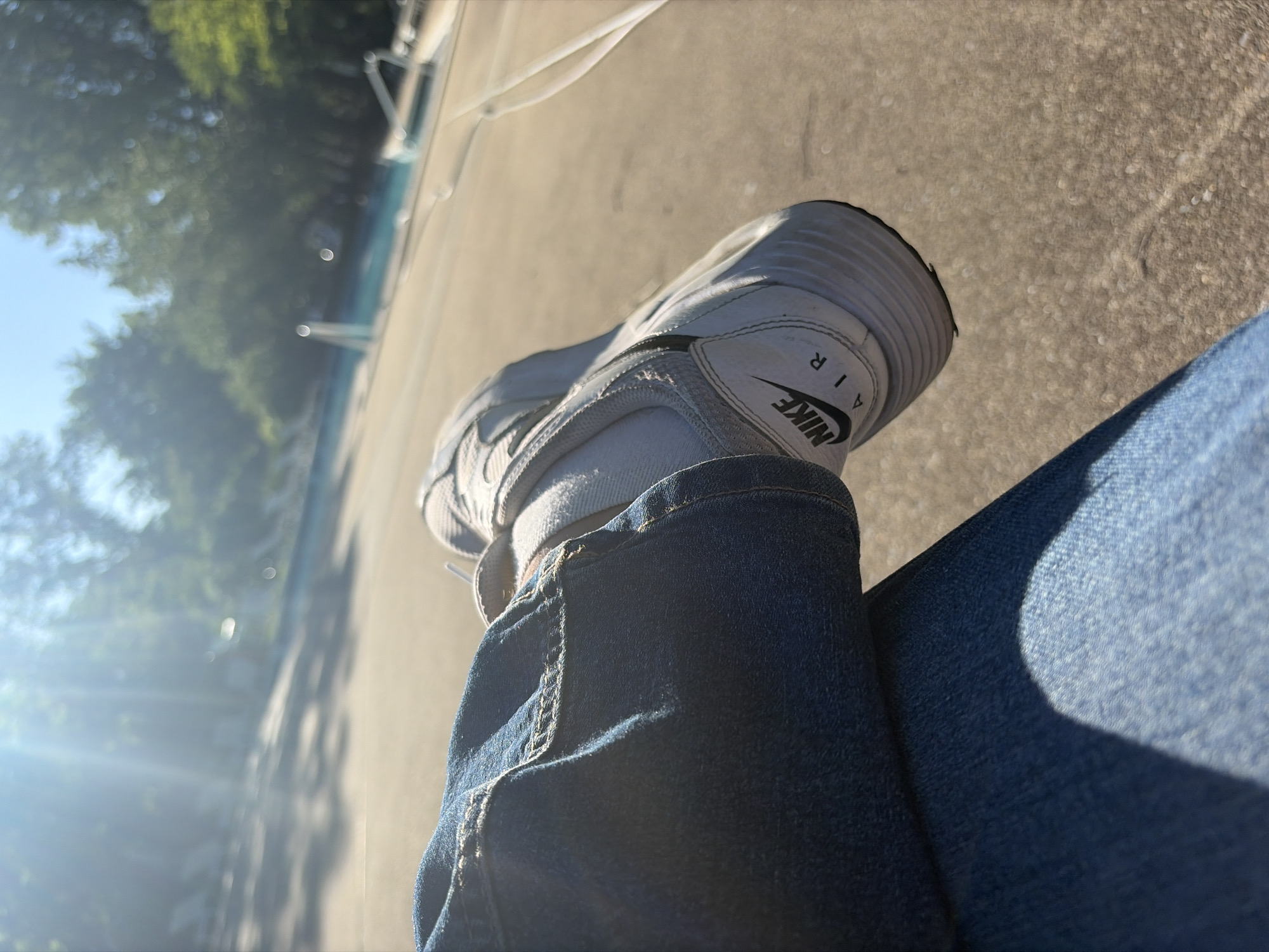 A close-up, angled shot focuses on a person's left leg clad in blue jeans and a white Nike Air sneaker, resting on a concrete surface, presumably outdoors. The jeans are denim with visible stitching and a pocket hem. The shoe exhibits the Nike logo. The background is blurred, showing trees and a sunlit area, possibly near a body of water. While the general location seems to be outdoors in a place with trees and concrete, there is insufficient data to determine the specific location in Edison, United States of America.