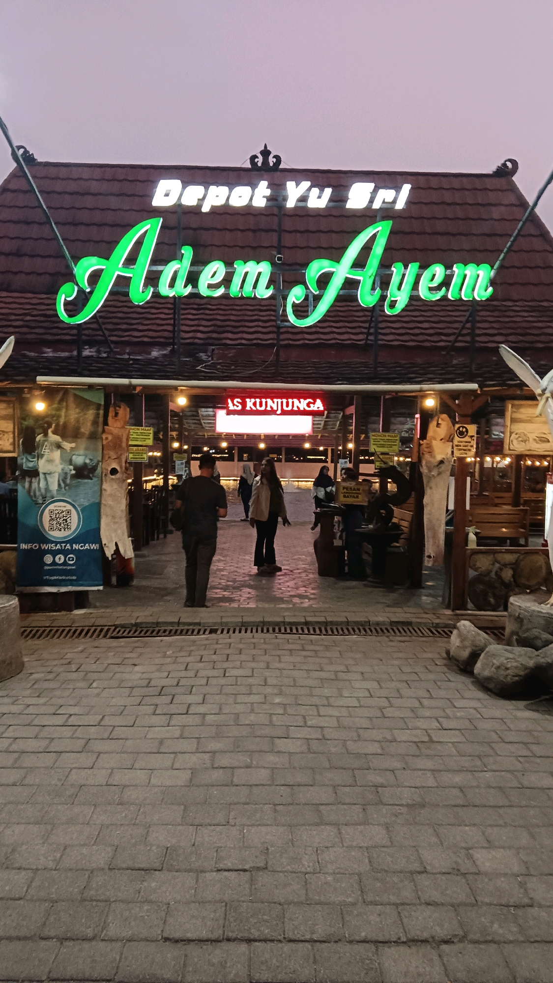 The image depicts the entrance to a restaurant named "Depot Yu Sri Adem Ayem" in Soka, Indonesia. The name of the establishment is prominently displayed in illuminated green neon lettering against a brown tiled roof structure. Above the green lettering, "Depot Yu Sri" is written in white illuminated letters. Below the main signage, a red illuminated sign reads "AS KUNJUNGAN".
The scene shows a paved walkway leading into the restaurant. Several people are visible in the mid-ground. To the left, a banner with an image of people and animals and a QR code is displayed. The environment appears to be an outdoor setting, possibly a dining area with tables and benches visible within the establishment. The lighting suggests it is dusk or evening. The overall impression is that of a casual dining establishment.