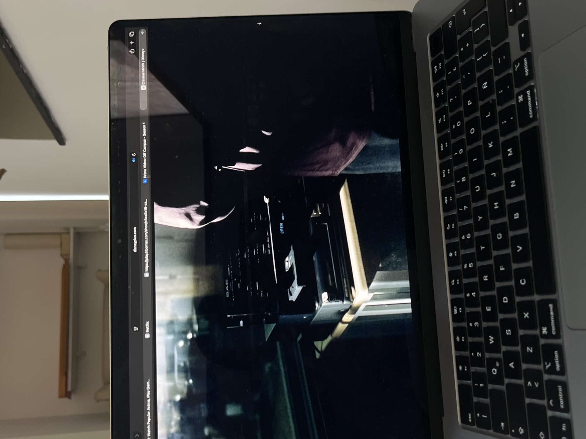 A laptop displays a scene from a television show, possibly "Criminal Minds" on Disney+, with a dark, indoor setting featuring audio equipment. The laptop is placed on a table in a room. The background shows a shelf with items on it, suggesting a domestic or office environment. The keyboard of the laptop is visible in the foreground. The scene is captured from a slightly high angle, looking down at the laptop.