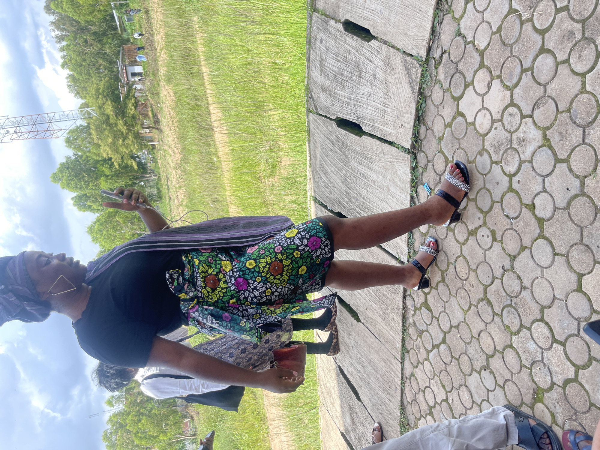A group of people are standing on a paved area with a textured, circular pattern. A woman in the foreground is wearing a black t-shirt, a colorful floral skirt, and a purple and white striped scarf draped over her shoulder. She is also wearing black sandals with a decorative strap. She is holding a mobile phone in her right hand. Behind her, another woman in a white shirt and patterned skirt is visible, carrying a dark bag. In the background, there is a grassy field, trees, and a communication tower. The ground is made of concrete slabs with gaps between them. The scene appears to be outdoors, under a cloudy sky. The location is identified as the city of Jos, Nigeria.