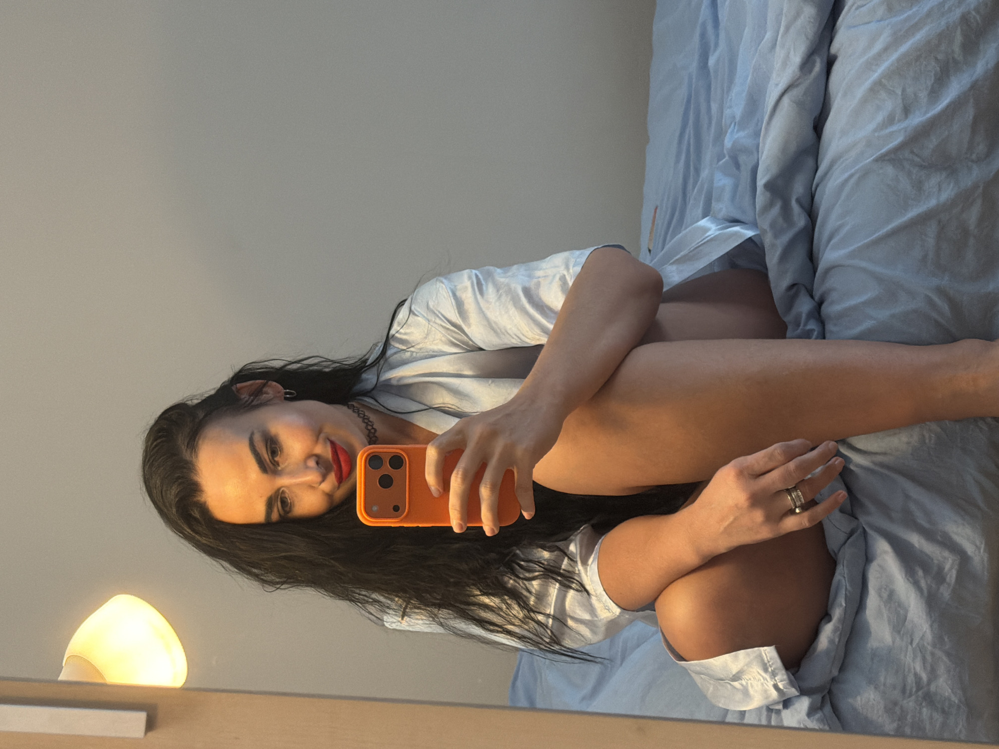 A woman with dark hair and red lipstick is lying in bed, taking a selfie with an orange smartphone. She is wearing a light-colored satin shirt. The bed has light blue sheets. A lamp is visible in the lower left corner of the image. The location is Unknown, Unknown.