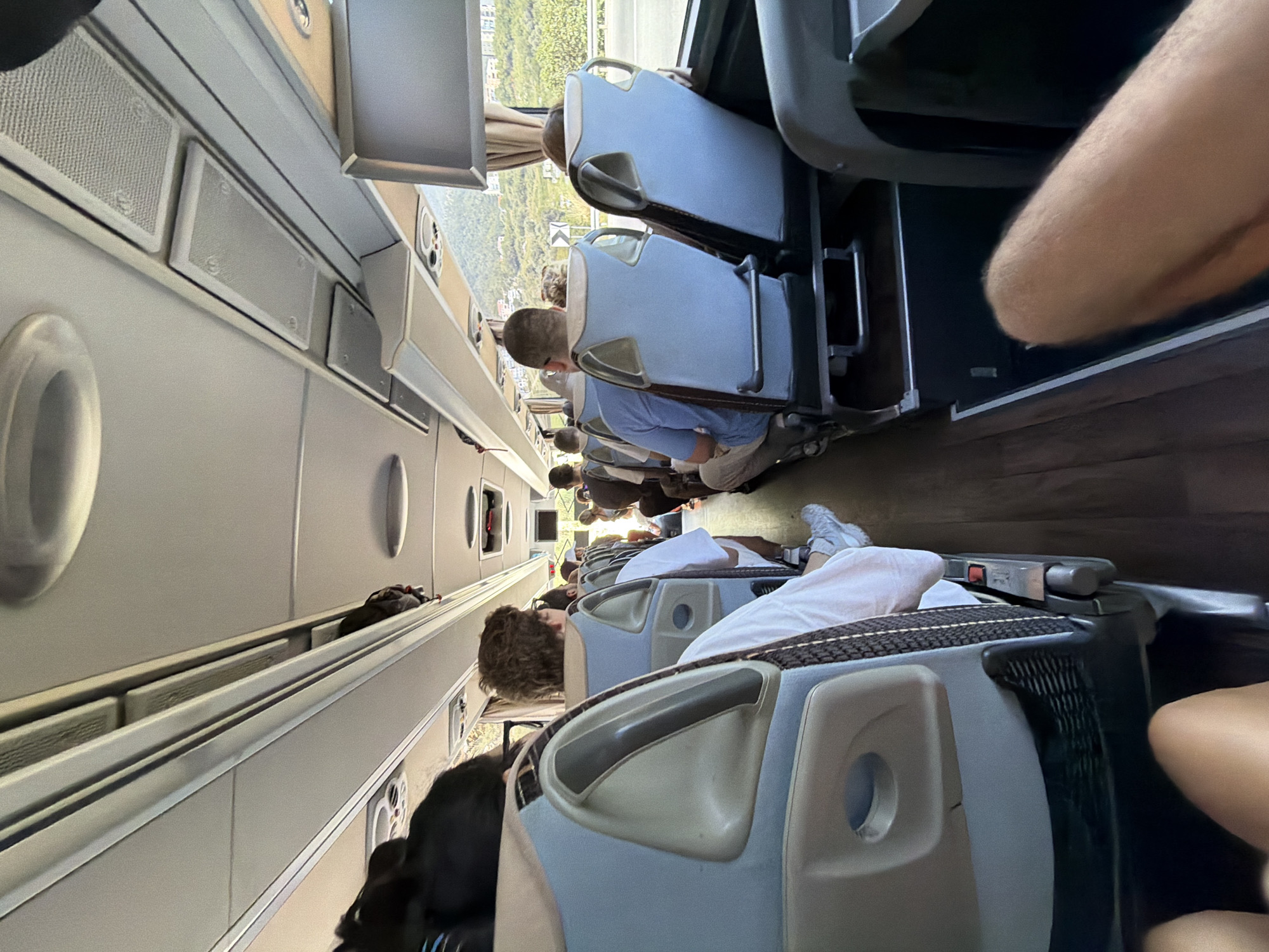 The image depicts the interior of a passenger bus, viewed from a low angle looking forward down the aisle. Several passengers are seated in the blue and grey upholstered seats, facing forward. The aisle is lined with wooden flooring. Overhead storage compartments are visible along the ceiling, with bags stored in them. A window on the right side of the bus shows a view of a hillside with buildings and vegetation, suggesting a location near a populated area. The lighting appears to be natural, coming from the windows. The scene is static, with no discernible action taking place. The overall impression is of a typical bus journey. This scene is located in Donji Pobori, Montenegro.