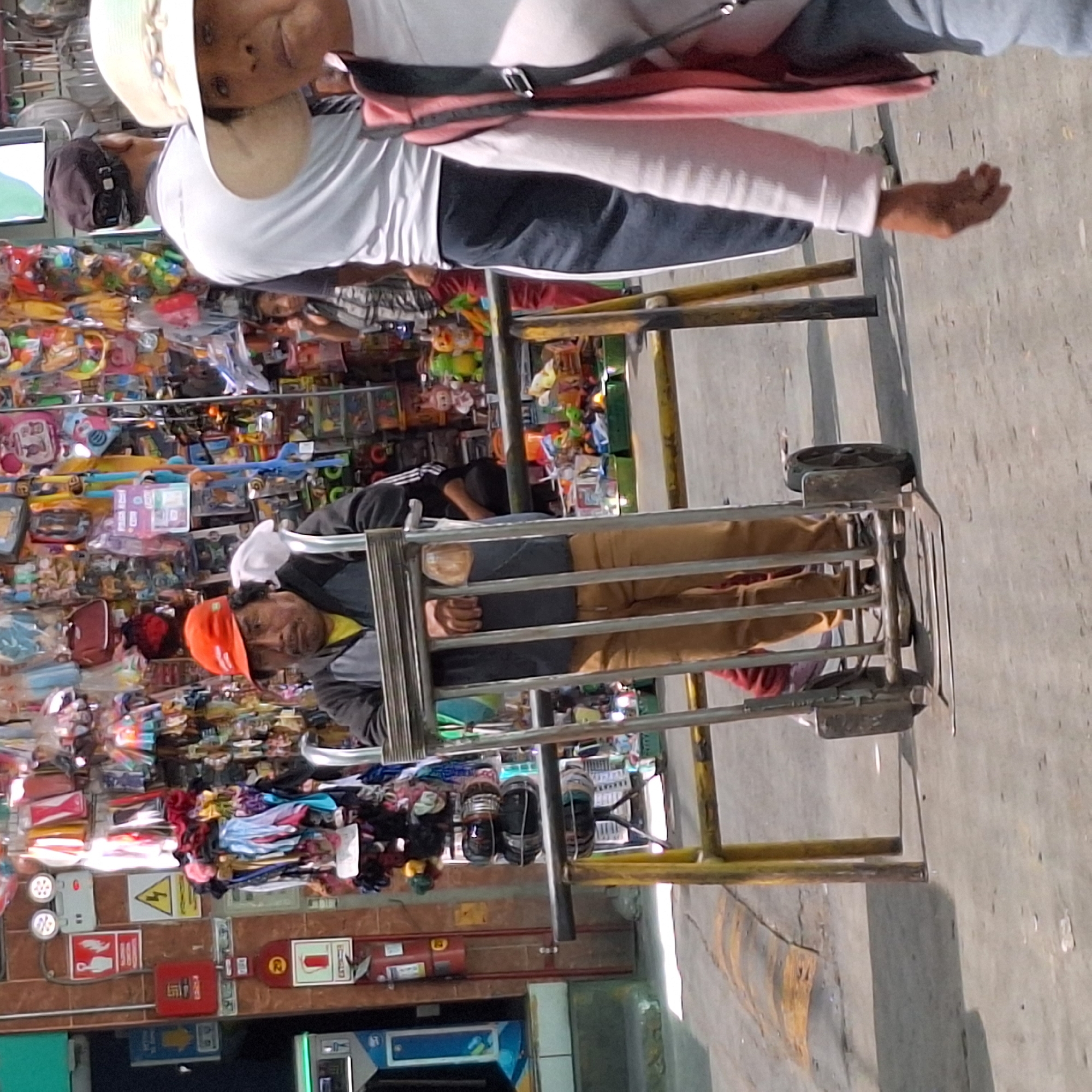 This image depicts an outdoor market scene in José Luis Bustamante y Rivero, Peru, under bright daylight conditions.
In the upper portion of the frame, a person with light skin wears a white long-sleeved shirt, dark pants, and a light-colored hat, with a pink/red bag strapped across their body. Their face is visible, looking towards the left. Partially obscured behind them is another individual in a light grey shirt and a dark cap.
In the lower center, a market vendor is seated within a metal-framed cart or stall. This person wears an orange baseball cap, a dark grey jacket over a yellow shirt, and light brown pants, looking to the right. The cart, equipped with wheels, suggests mobility.
The background is filled with numerous market stalls displaying a dense assortment of colorful merchandise, including what appears to be toys, clothing, and various small goods, typical of a vibrant street market. The ground is a light-colored concrete or pavement.
To the left, a brown tiled wall features several affixed signs, including a yellow triangular warning sign, red square signs (one with a white symbol, possibly fire-related), and a visible red fire extinguisher. A blue and white machine, resembling an ATM, is also mounted on the wall further down. The overall scene portrays a busy day of commerce in a public outdoor setting. No specific text on the signs is clearly legible beyond their general symbols.