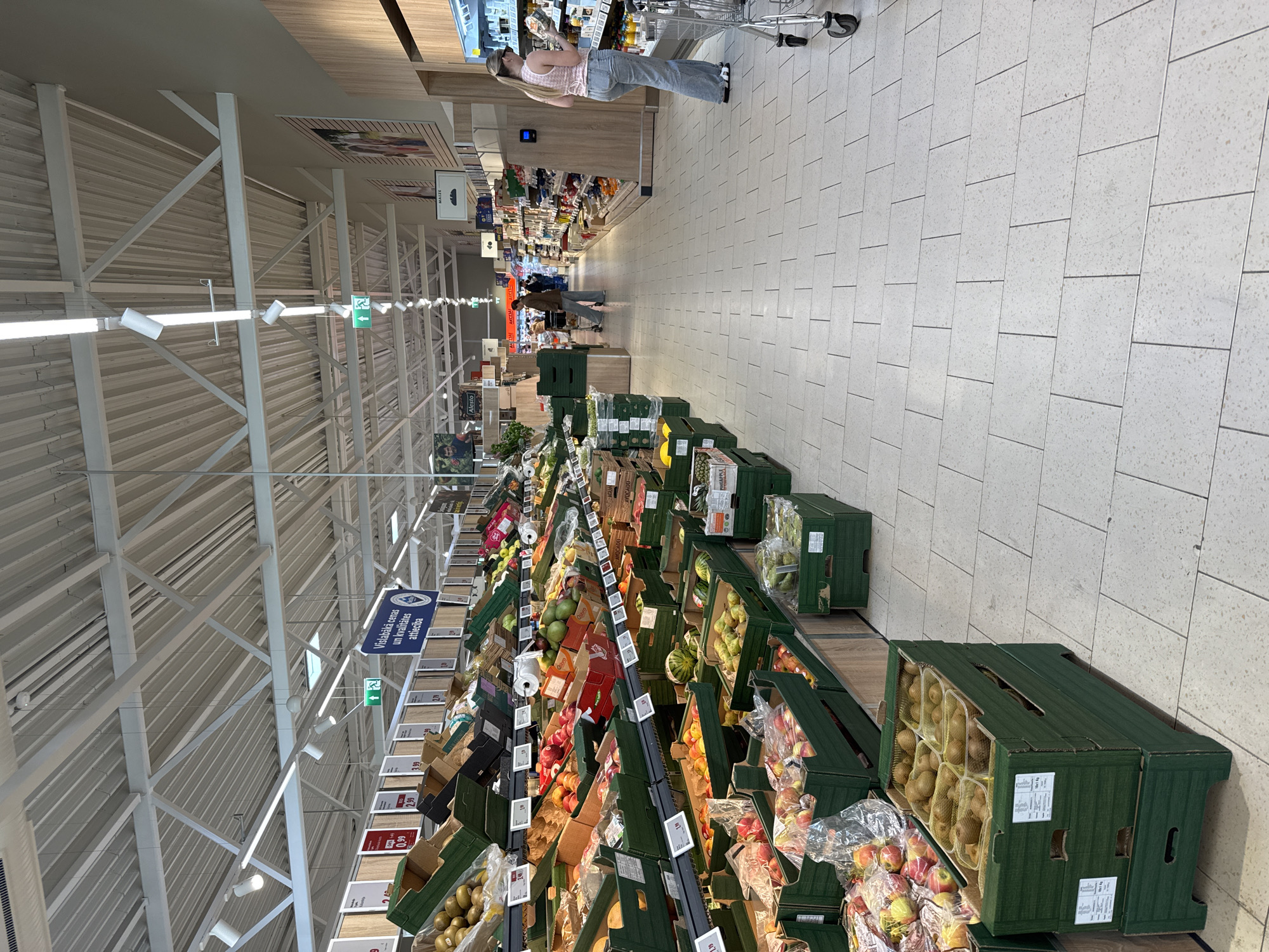 The image depicts an aisle in a supermarket, possibly in Riga, Latvia, based on the text visible on some labels. The focus is on the fruit and vegetable section, with numerous boxes of produce arranged along the aisle.
Several individuals are visible, including a woman in a pink top and jeans pushing a shopping cart at the far end of the aisle. Other individuals can be seen further down the aisle, examining the produce.
The environment is a typical supermarket interior, with fluorescent lighting, a high ceiling with metal supports, and tiled floors. The boxes of fruits and vegetables are stacked in green containers. Signs with prices and product information are visible in the aisle.