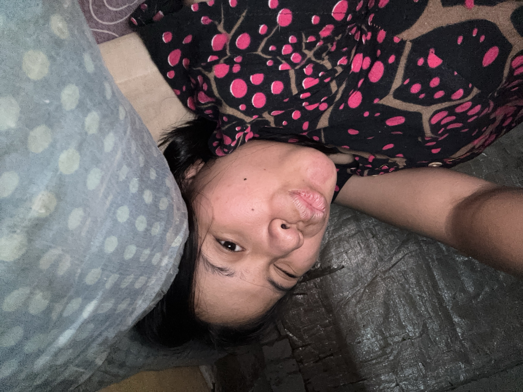 A woman is lying down and making a kissy face at the camera, with one eye winking. She is wearing a black top with a pattern of pink polka dots and brown branches. She is lying on a surface that appears to be a tarp, and a pillow with a light blue and white polka dot pattern is next to her. The scene is dimly lit, suggesting it is indoors or nighttime. The location is East Jakarta, Indonesia.