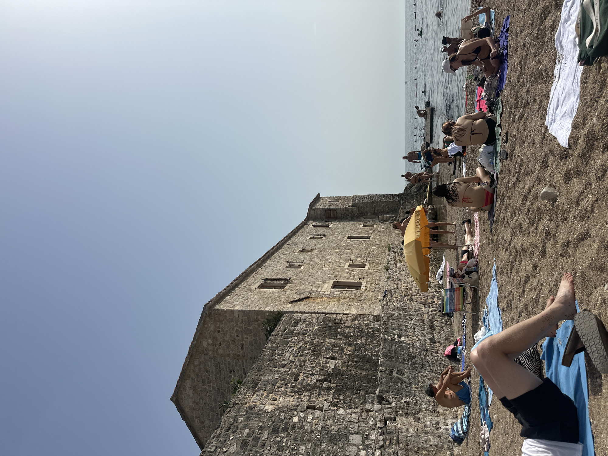 The image depicts a sunny day at a beach adjacent to a historic stone building, likely within the old town of Budva, Montenegro. The building, constructed from rough-hewn stone, features small windows with wooden shutters and appears to be a significant historical structure. On the sandy beach in front of the building, several people are sunbathing and relaxing on towels. Some individuals are seated, while others are lying down. A yellow umbrella provides shade for a group of people. The sea is visible in the background, with several people swimming or wading in the water, and small boats can be seen in the distance. The overall atmosphere is one of a leisure activity on a warm day.