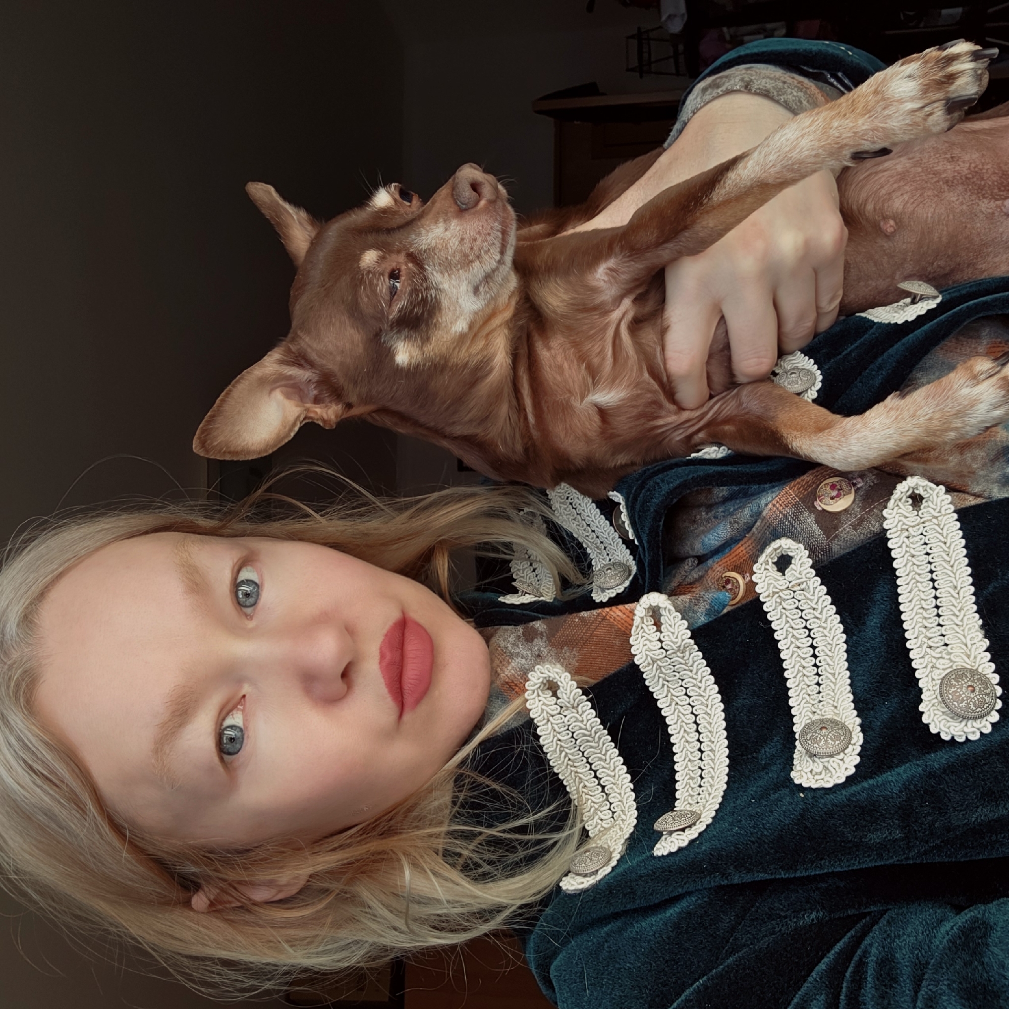 A person with light skin, blue eyes, and blonde hair is holding a brown Chihuahua. The person is wearing a dark green velvet jacket with white decorative trim and silver buttons. The background is dark and indistinct. There is no visual indication of location, and therefore no way to determine if the photo was taken in Saint Petersburg, Russia.
