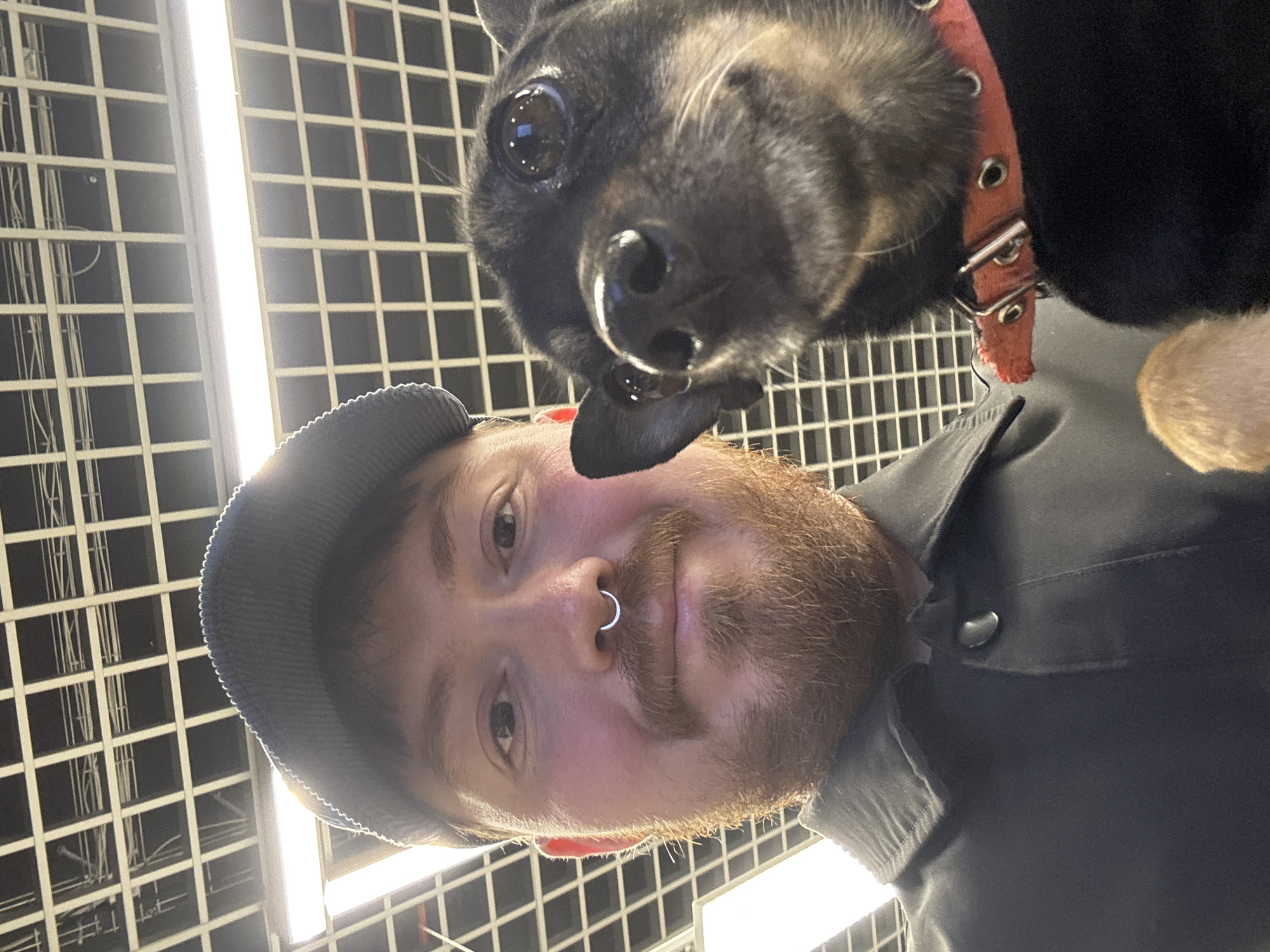A man and a dog are in an indoor setting. The man is wearing a dark cap and shirt with a nose ring. The dog is small, black and tan, and wearing a red collar. The background features a grid-like ceiling structure. The location cannot be confirmed as Chaikovsky, Russia without further information.
