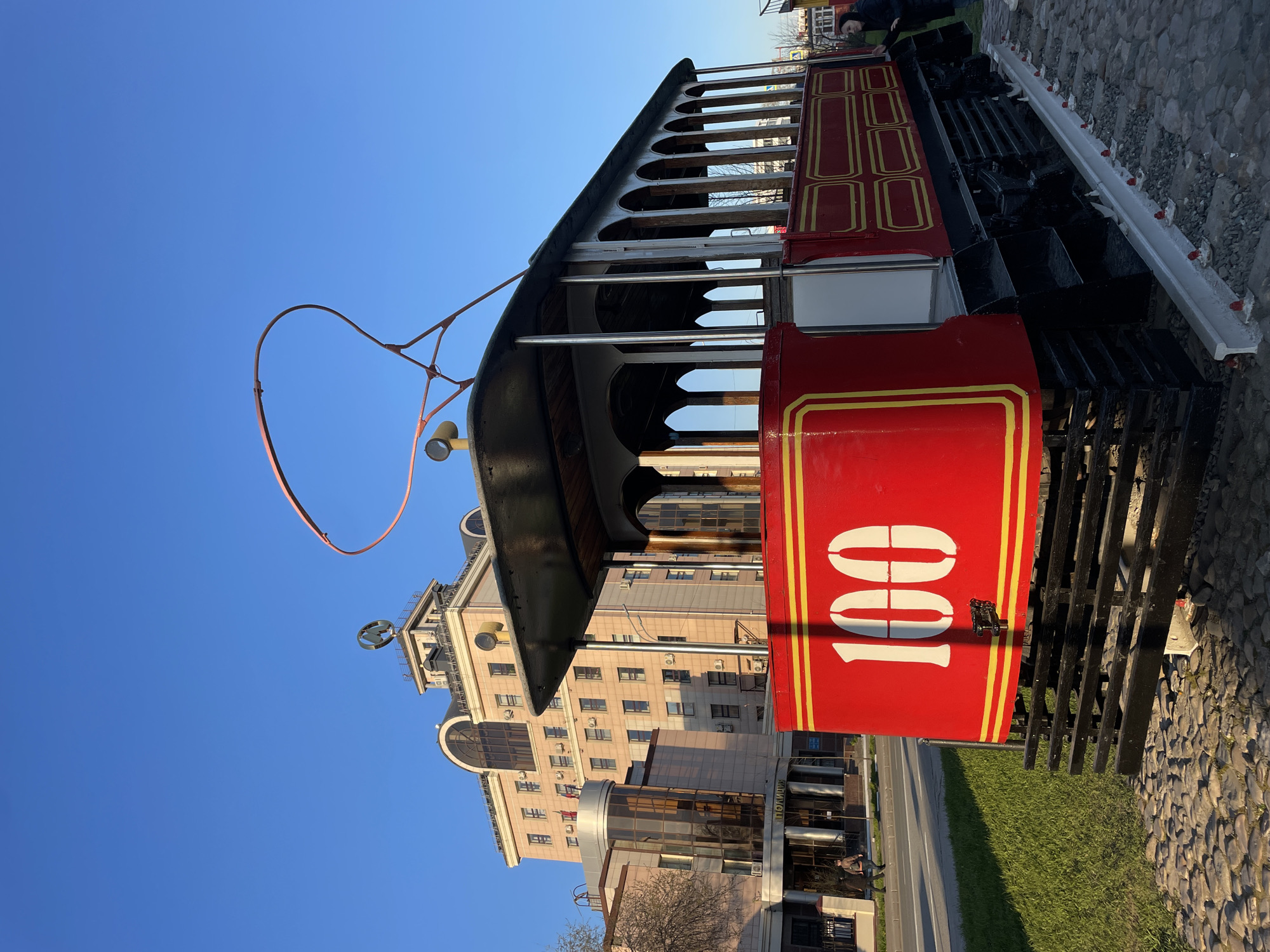 The image shows a red and gold trolley car with the number "100" prominently displayed on its side. The trolley is positioned on a stone and grass area with a building visible in the background. The building appears to be modern, multi-story, and likely commercial or office space. It has a large "W" logo. The sky is clear and blue, suggesting fair weather. The location is Kazan, Russia.