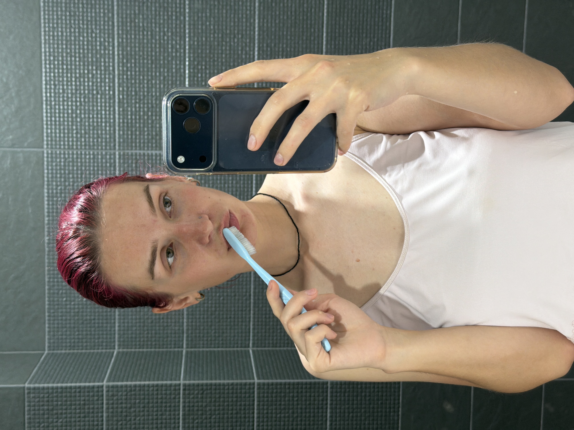 A young woman with dyed pink hair is taking a selfie in a bathroom. She is wearing a light pink camisole and has a black beaded necklace on. In her right hand, she holds a light blue toothbrush to her mouth, as if she is brushing her teeth. Her left hand holds a dark blue smartphone, angled to capture her reflection in the mirror. The bathroom has dark grey tiled walls. The image was taken in Chaikovsky, Russia.
