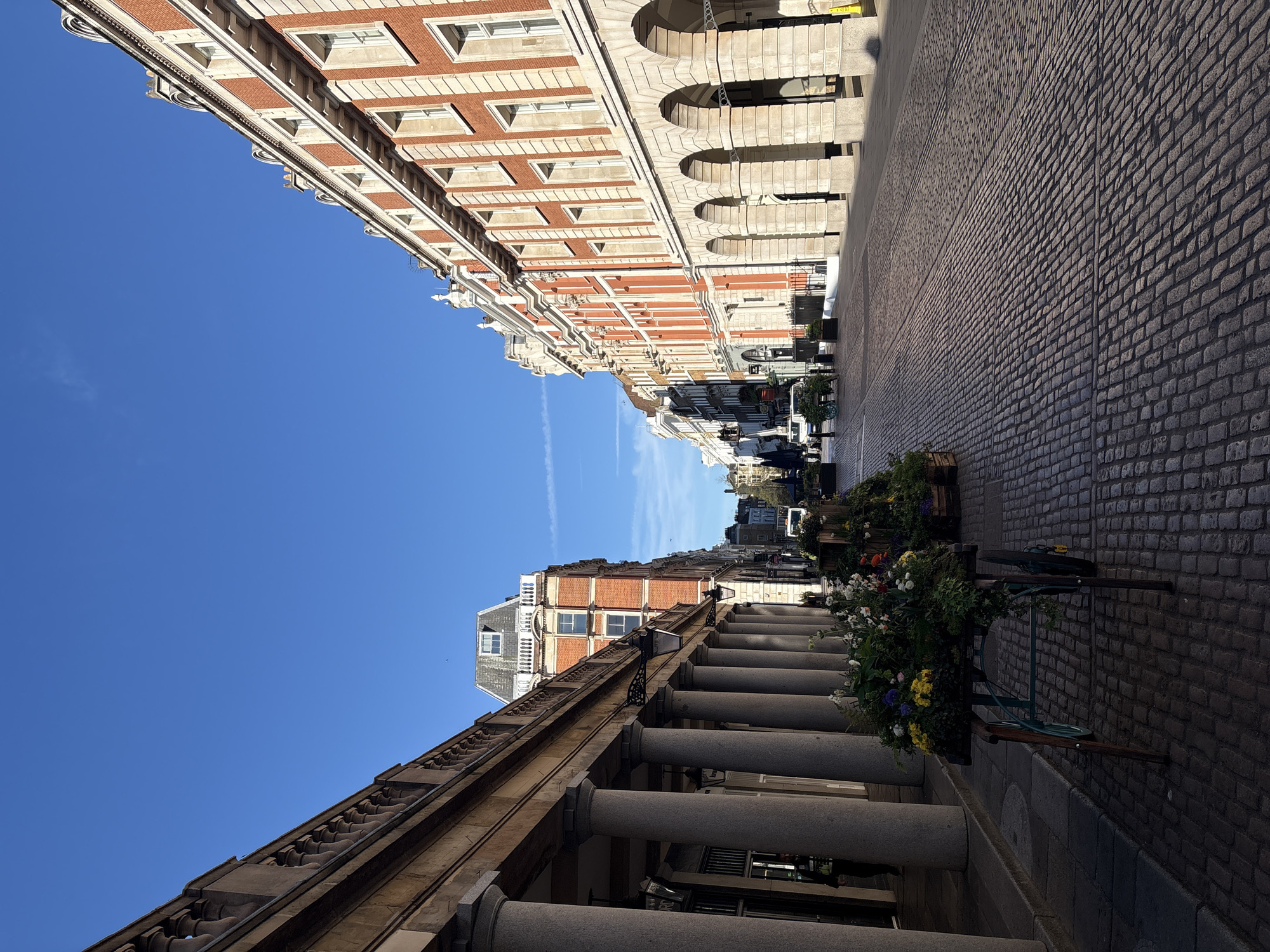 The image depicts a street in London, United Kingdom. The perspective is looking down a cobblestone street, flanked by buildings with distinctive architectural styles. The building on the left features a colonnade with large, cylindrical columns. The building on the right has a facade of red brick and white stone. The sky is clear blue. The scene is devoid of people. Flower baskets are visible along the sidewalk.