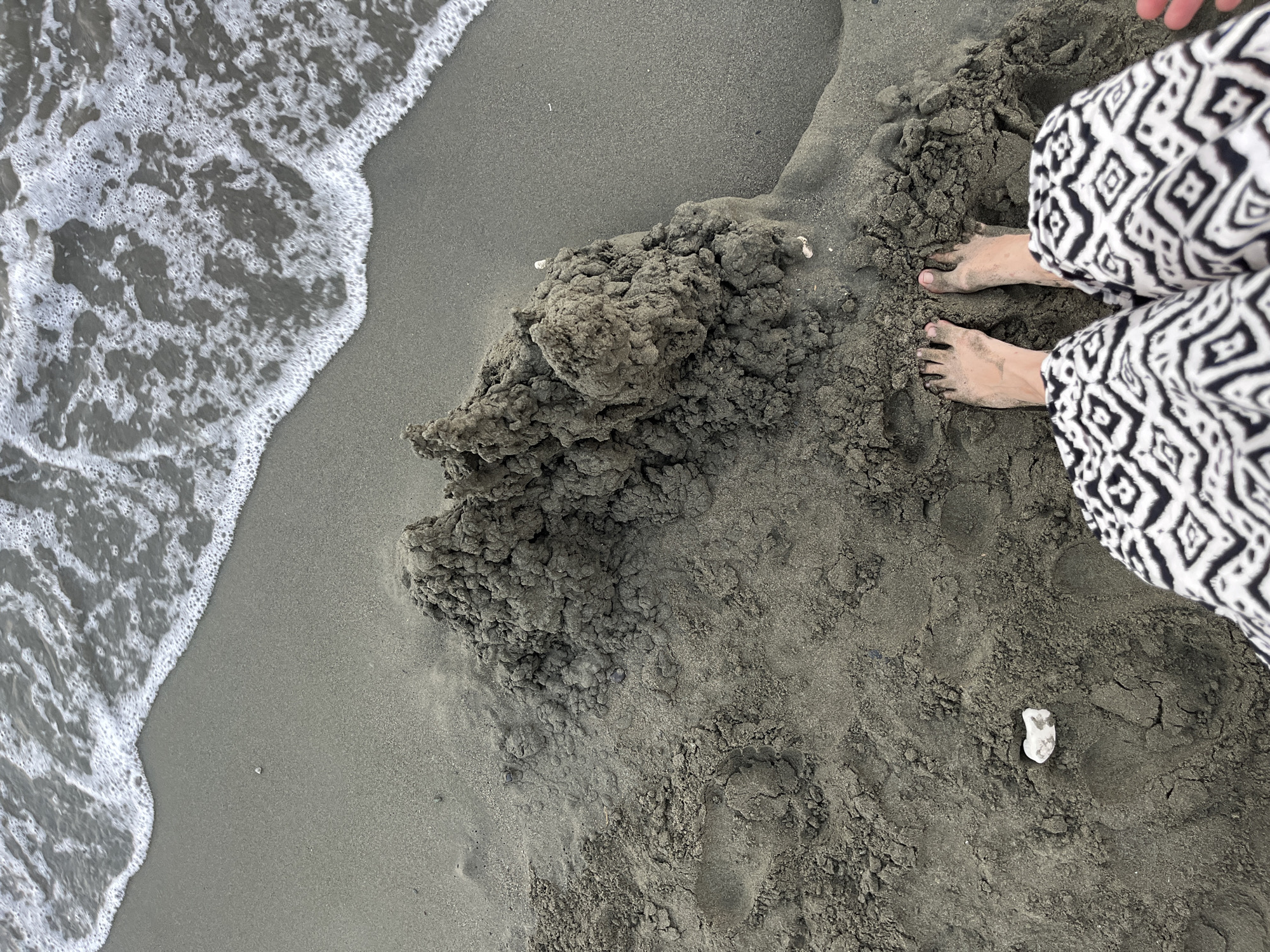 An overhead shot captures a person's bare feet standing on a dark, sandy beach. The person is wearing black and white patterned pants. To the left of the feet, a pile of dark, wet sandcastle ruins is visible. The ocean waves with white foam are encroaching on the sand from the top of the frame. A small white shell is present in the sand to the left of the sandcastle. The location is identified as Agios Tychona Community, Cyprus.