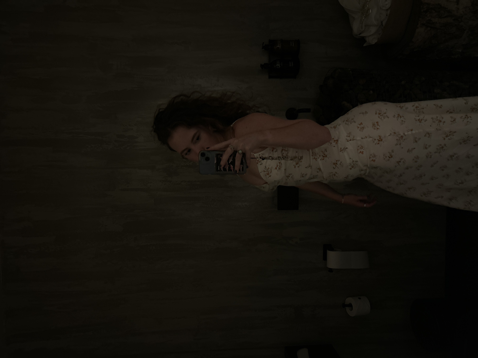 A dimly lit, rotated image depicts a female taking a mirror selfie in what appears to be an indoor setting, likely a bathroom, given the visible toilet paper dispenser. The woman, with dark, wavy hair, is wearing a light-colored, possibly white or cream-colored, floral dress featuring small orange or brown blossoms. She holds a dark-colored smartphone in her left hand, which is adorned with rings and dark nail polish. Her face is partially visible in the reflection as she looks towards the phone.
The background features dark, textured walls, possibly concrete or a dark tile, contributing to the overall dim ambiance. In the upper right corner, two dark-colored pump bottles, likely for toiletries, are visible on a ledge or shelf. Below the person's torso, a white toilet paper dispenser with a roll of toilet paper is mounted on the wall. Above the person's head, in the reflection, a white, ornate decorative item, possibly a statue, is visible, alongside some dark, rocky, or textured material. The lighting is very low, making many details indistinct. There is some illegible white text on the dark bottles in the background. The image is taken in a casual moment, consistent with a selfie. The external context indicates the photo was taken in Krasnoyarsk, Russia.