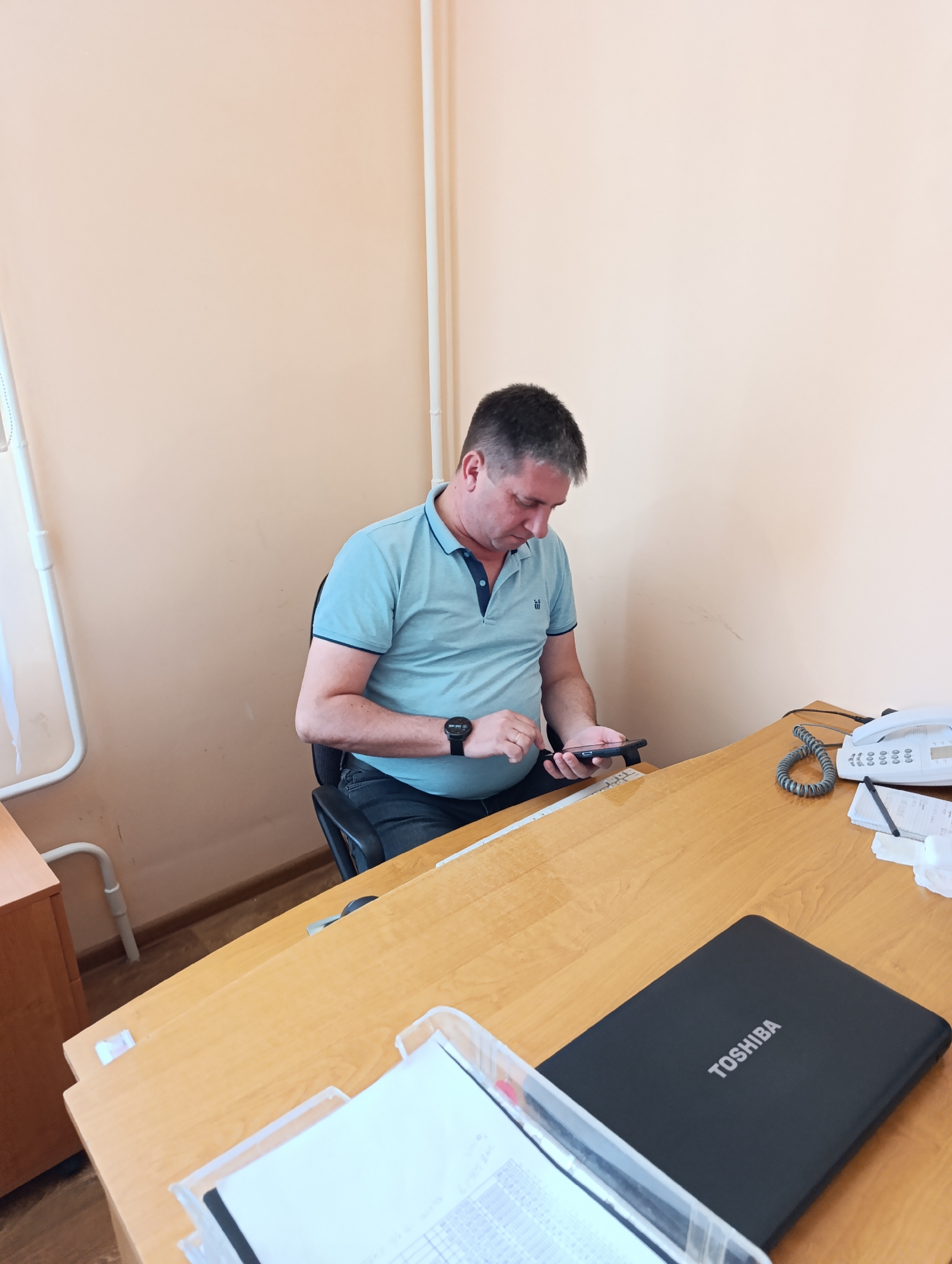 A man is seated at a wooden desk in an office, looking down at his smartphone. He is wearing a light blue polo shirt and dark jeans. On the desk, there is a white corded phone, a black laptop with the Toshiba logo, a white paper in a clear plastic holder, and a ruler. The office walls are painted a light peach color. The scene appears to be within the Dubăsari town council building in Moldova.