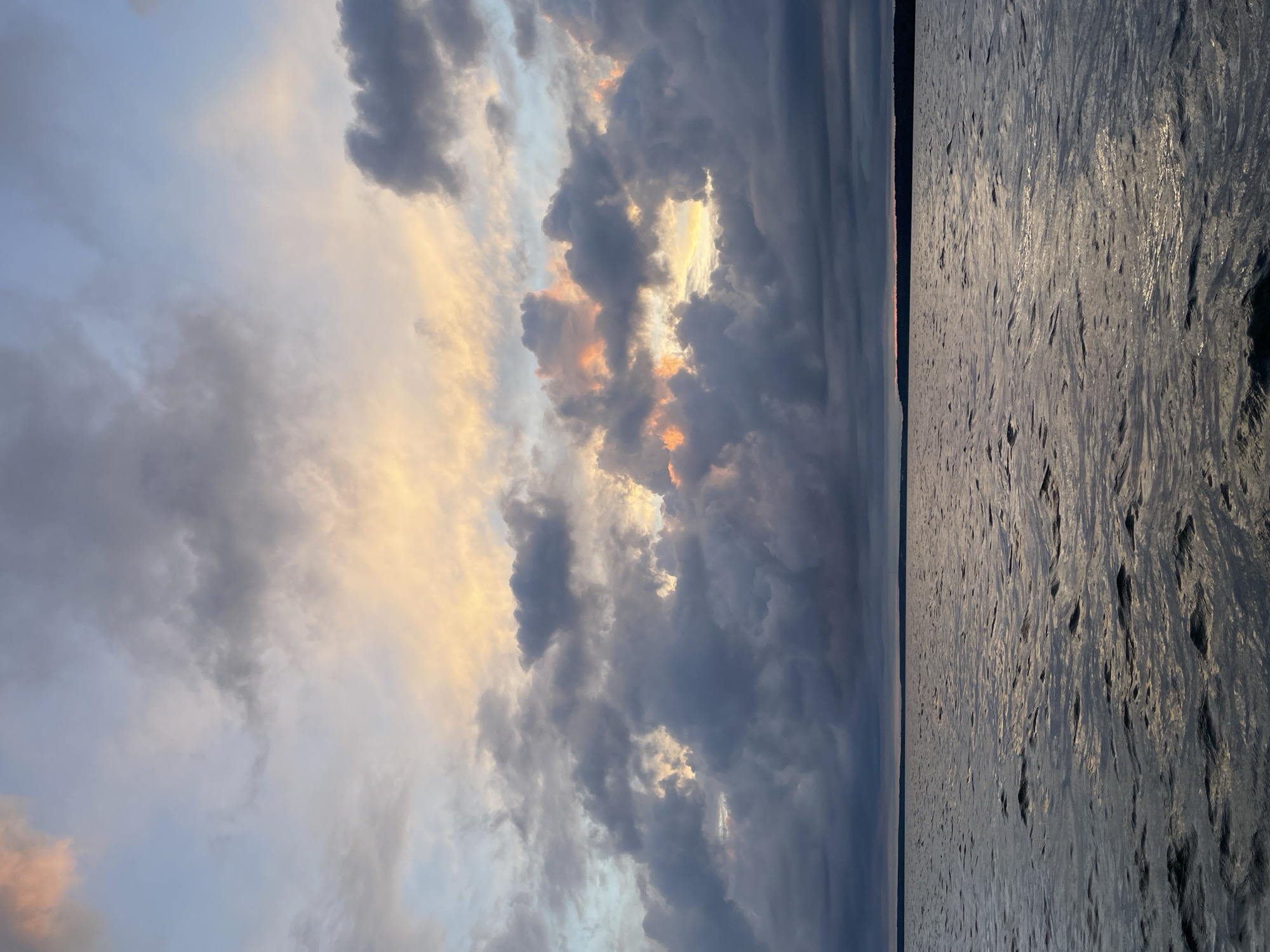 A vast body of water stretches towards a distant, dark horizon line, indicating land. The water's surface is slightly rippled, catching and reflecting the muted, warm light from the sky above. The sky is filled with dramatic, heavy clouds in various shades of grey, with hints of soft pink and golden light breaking through in the upper right quadrant. The overall atmosphere is serene and somewhat dramatic, evoking a sense of approaching dusk. There are no people, specific structures, or distinguishing landmarks visible in this image, making it impossible to determine a specific location such as Yablonovka, Russia.