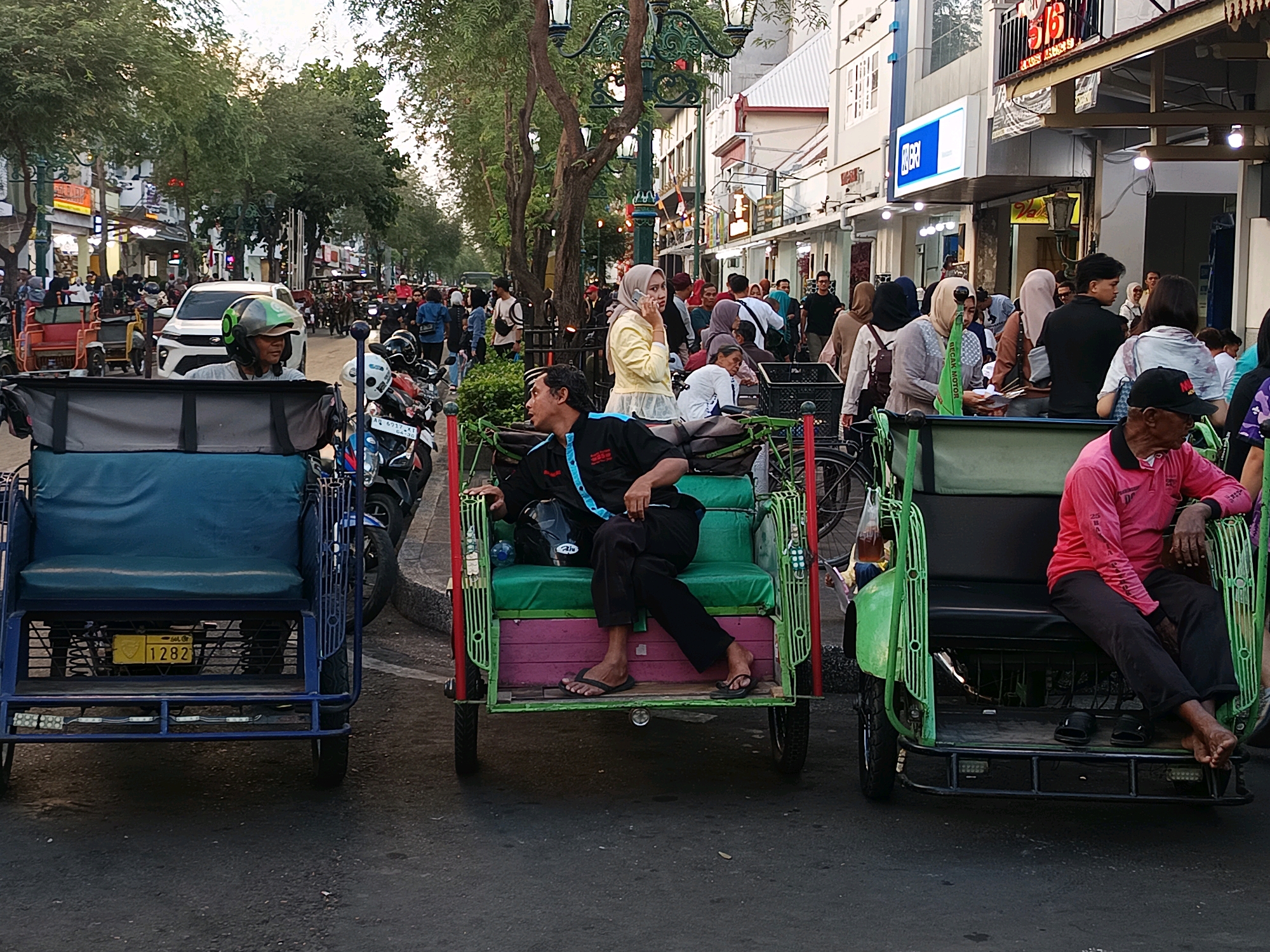 The image depicts a street scene in Yogyakarta, Indonesia, likely Malioboro Street, known for its shops and street vendors. The foreground features three rickshaws, also known as becak. The rickshaw on the left is blue and empty. The middle rickshaw is green and pink, occupied by a man in a black shirt and pants, wearing flip-flops. The rickshaw on the right is green and occupied by a man in a pink long-sleeved shirt and black pants, also wearing flip-flops.
In the background, a white car is visible, along with various motorcycles. A crowd of people, dressed in casual attire, are walking along the sidewalk and the street. Some individuals are wearing headscarves. Buildings line the street, many of them appearing to be shops with signage. Trees with lush green foliage contribute to the urban landscape. The overall atmosphere suggests a busy, bustling street during daylight hours, likely late afternoon or early evening given the lighting.
