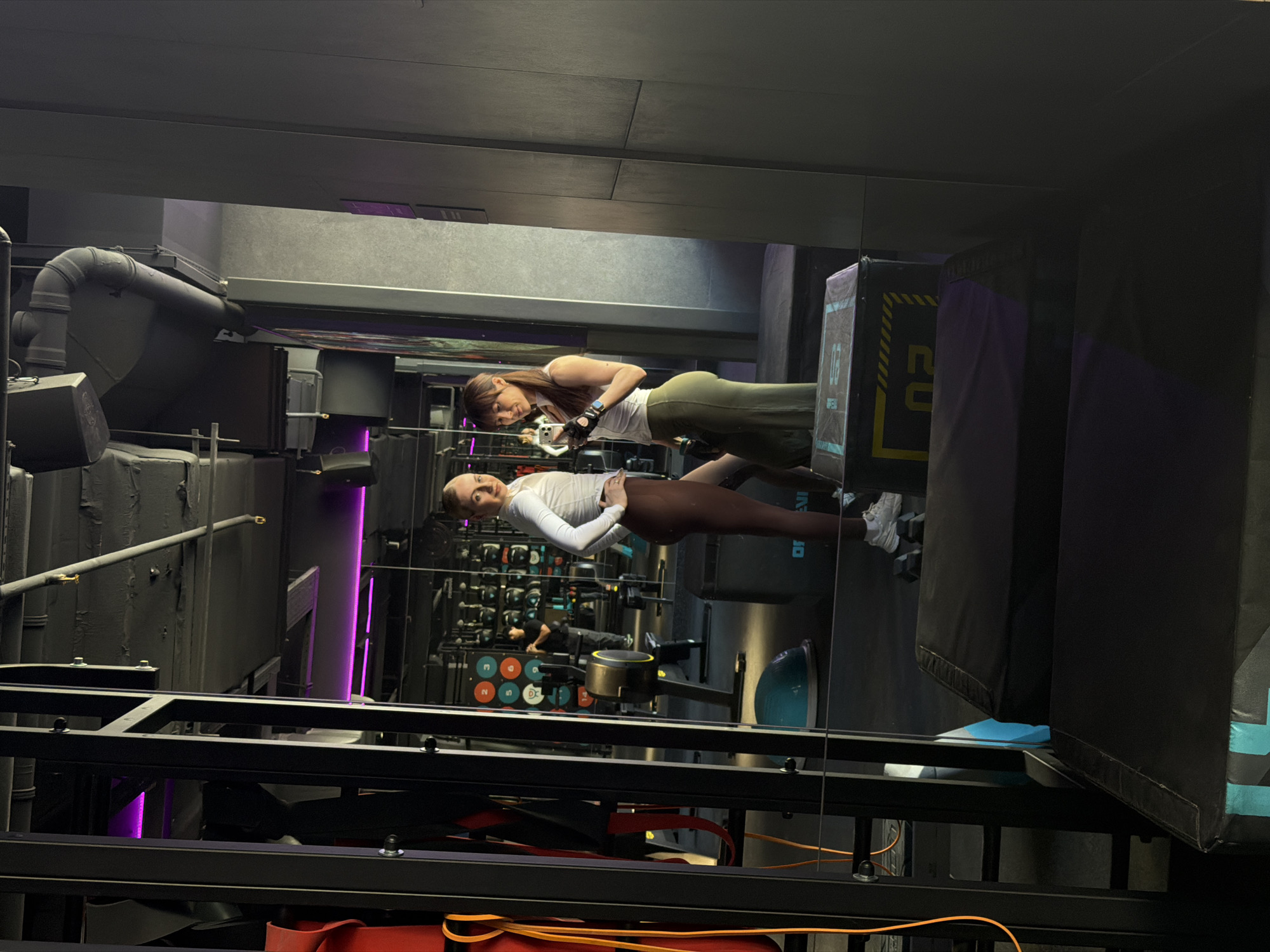 The image depicts the reflection of two women in what appears to be a gym or fitness center, possibly located in Perm, Russia, given the presence of Russian text on equipment and the overall gym style. The women seem to be taking a selfie or recording a video, with one holding a phone. The gym environment is characterized by industrial-style elements with exposed pipes, black framing, and workout equipment. Color-coded weights, machines, and soft blocks can also be seen. The gym's equipment and layout suggest a functional fitness or crossfit-type training facility.