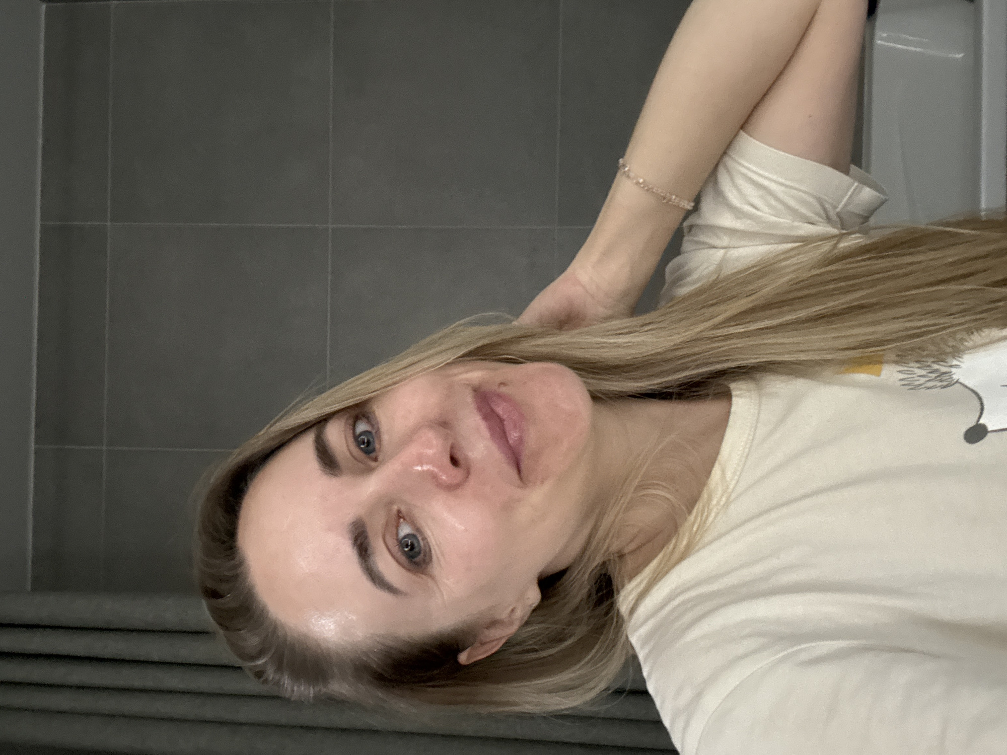 A light-skinned woman with blonde hair and blue eyes is posing for a selfie in what appears to be a bathroom. She is wearing a light-colored t-shirt and a beaded bracelet. The background features gray tiled walls and a gray-striped surface, possibly part of a vanity or wall covering. The specific location in Chaikovsky, Russia cannot be determined from the image.