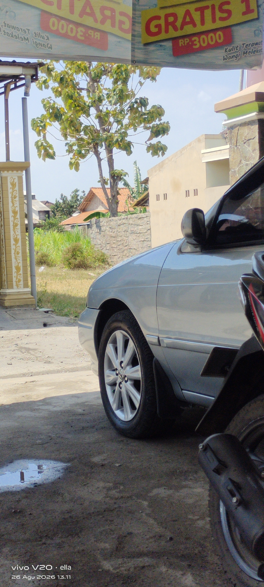The image shows a close-up view of a silver car parked on a dirt or gravel surface. The car's front fender and wheel are prominently visible. In the background, there is a tree with green leaves, a stone wall, and a building. A portion of a motorcycle is also visible in the foreground, to the right of the car. The sky is blue and clear. A sign in the background has text in yellow and red, including "GRATIS 1" and "RP.3000". The text "Kulit Terawat Wangi Melekat" is also visible on the sign. At the bottom of the image, there is a timestamp and text indicating the photo was taken with a "vivo V20 · ella" device on "26 Agu 2026 13.11". The scene appears to be outdoors, possibly in a residential or commercial area in Simo, Indonesia, given the presence of typical Indonesian architecture and vegetation. There are no people or animals visible in the frame.