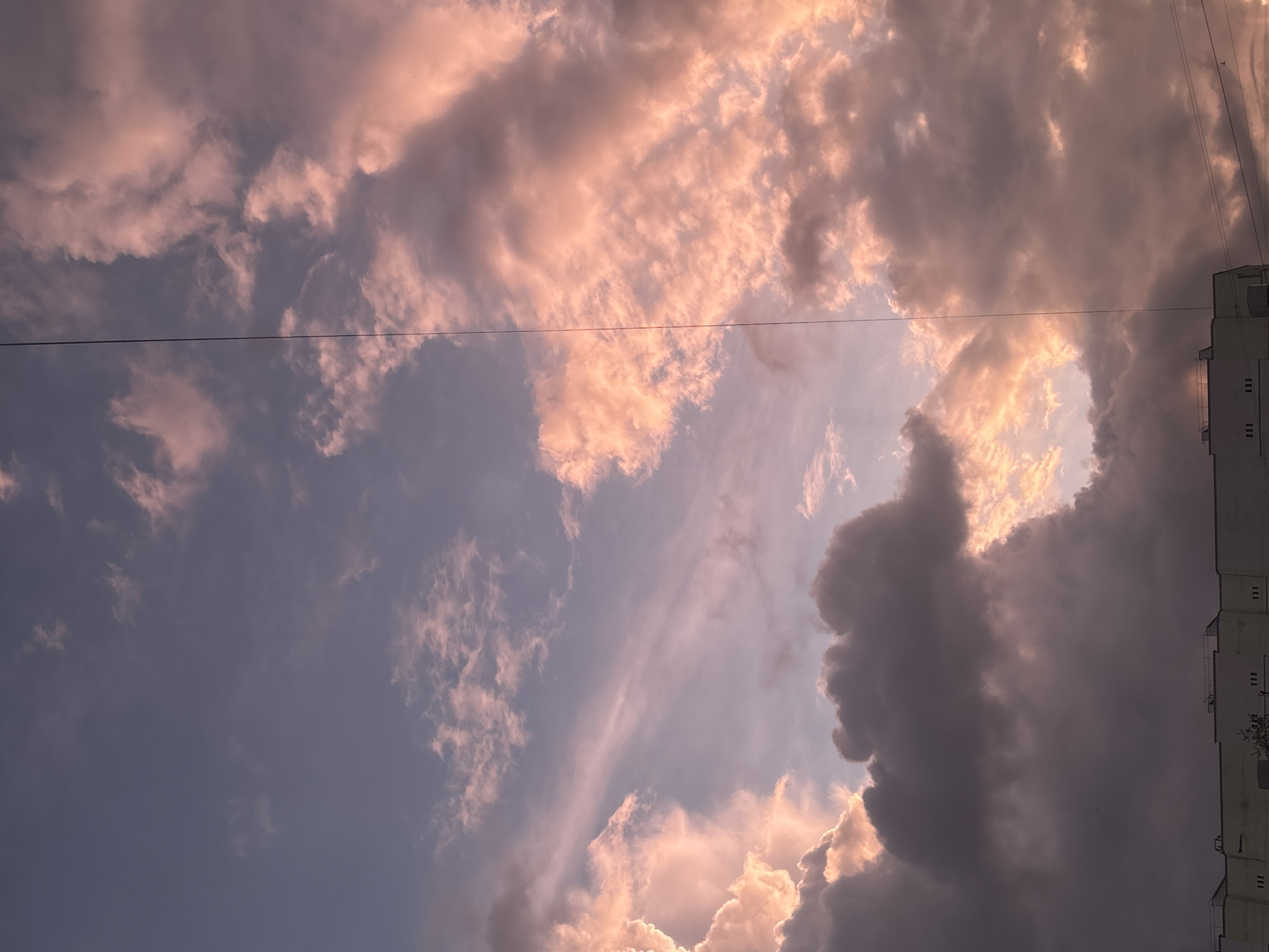 The image captures a dramatic sky filled with clouds, likely during sunset or sunrise. The sky transitions from a soft blue in the lower left to a more intense light towards the upper right, where the sun's warm glow is evident. Clouds vary significantly in appearance; some are illuminated by golden and pink light, showcasing soft, ethereal textures, while others are dense and dark grey, casting deep shadows. A large, dark cloud formation dominates the lower right portion of the frame, contrasting sharply with the brightly lit areas.
Two thin, dark power lines stretch horizontally across the upper mid-section of the image, breaking the vast expanse of the sky. On the far right, a segment of a multi-story building, light in color with visible windows, is partially visible, indicating an urban or suburban setting typical of places like Chaikovsky, Russia. No people or specific activities are depicted. The scene is a static observation of the interplay of light and cloud formations. There is no visible text in the image.