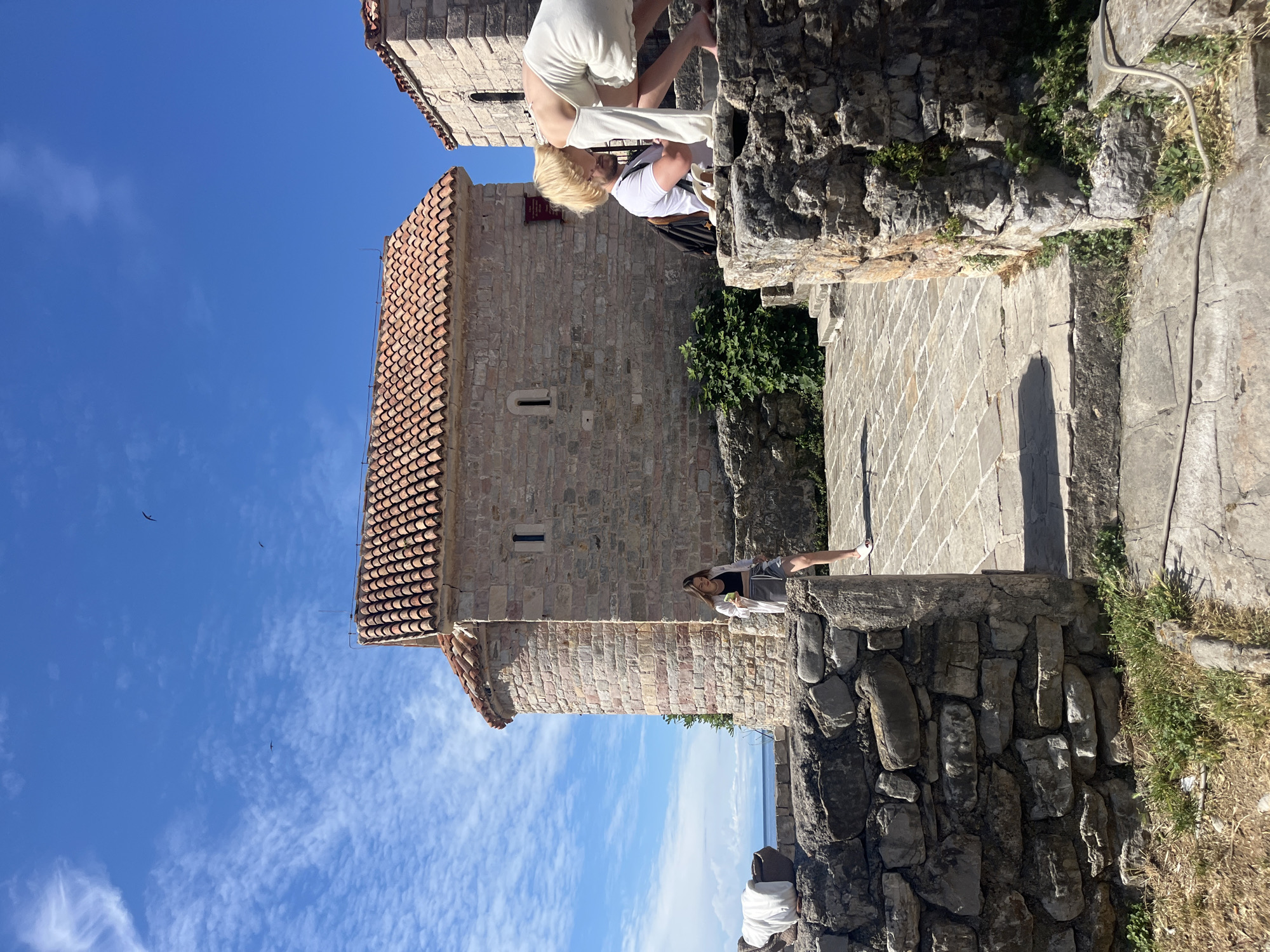In the city of Budva, Montenegro, at the Church of St. John, a circular stone building with a red tiled roof is visible under a bright blue sky with scattered white clouds. A woman with blonde hair, wearing a white dress, leans forward on a stone wall. To her left, a man with dark hair and a white t-shirt is seated. In the foreground, a stone wall divides the scene, with a path of stone pavers leading to the church. A young woman in a black top and grey shorts walks towards the church. In the background, the Adriatic Sea is visible.
