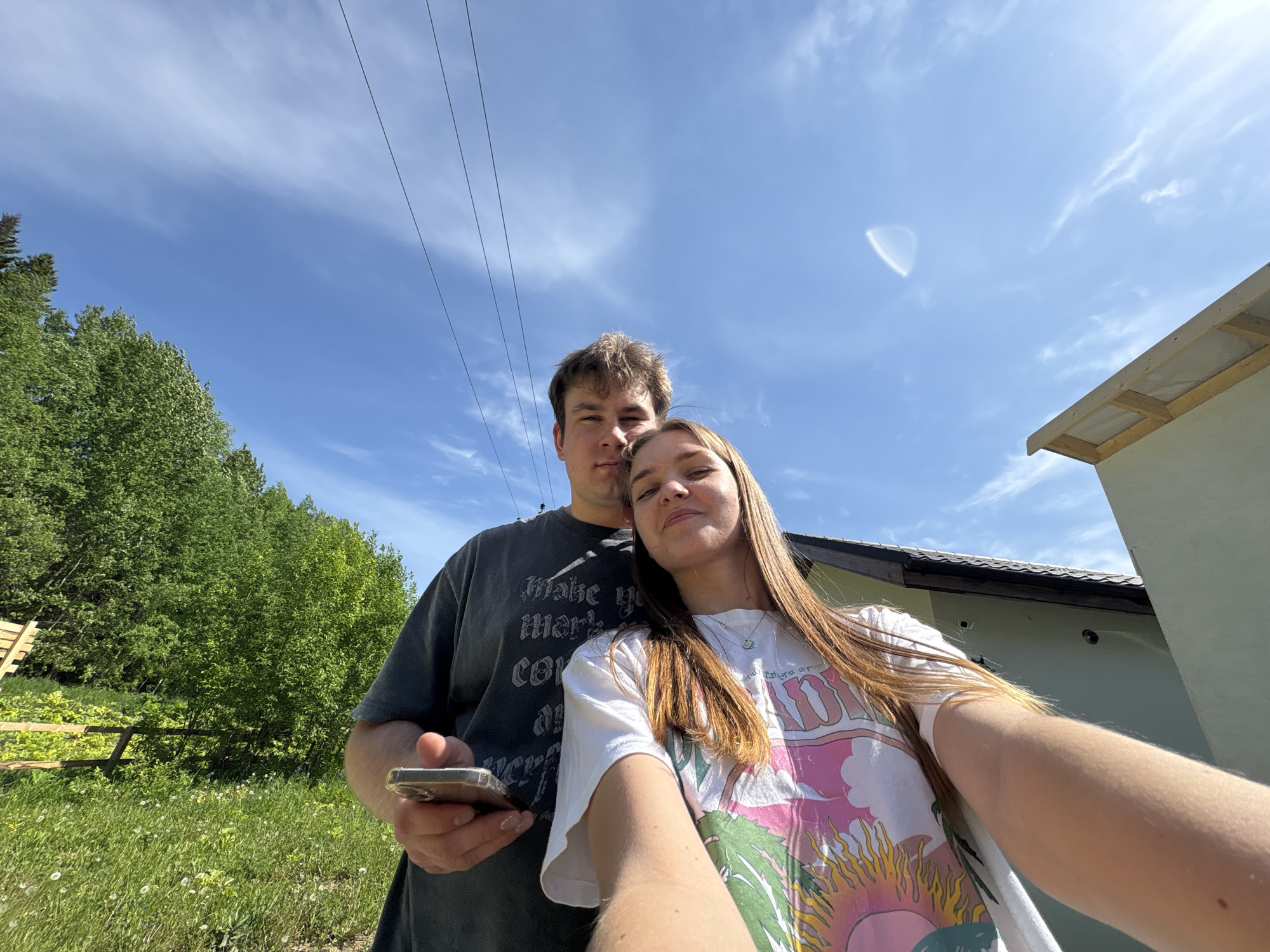 A medium shot shows a young couple standing close together, angled for a selfie, likely near a residential area in Sibir’, Russia. The woman, with long brown hair and wearing a white t-shirt with a colorful design, is holding the camera, extending her arm towards the viewer. A man stands behind her, wearing a grey t-shirt and holding a phone in his left hand. Behind them, there's a blue sky with light clouds and electrical lines. To the left, there is a grassy area with green trees and a partially visible wooden structure. To the right, there's part of a building with a light grey exterior. The sun is shining, as seen from the light on the subjects.