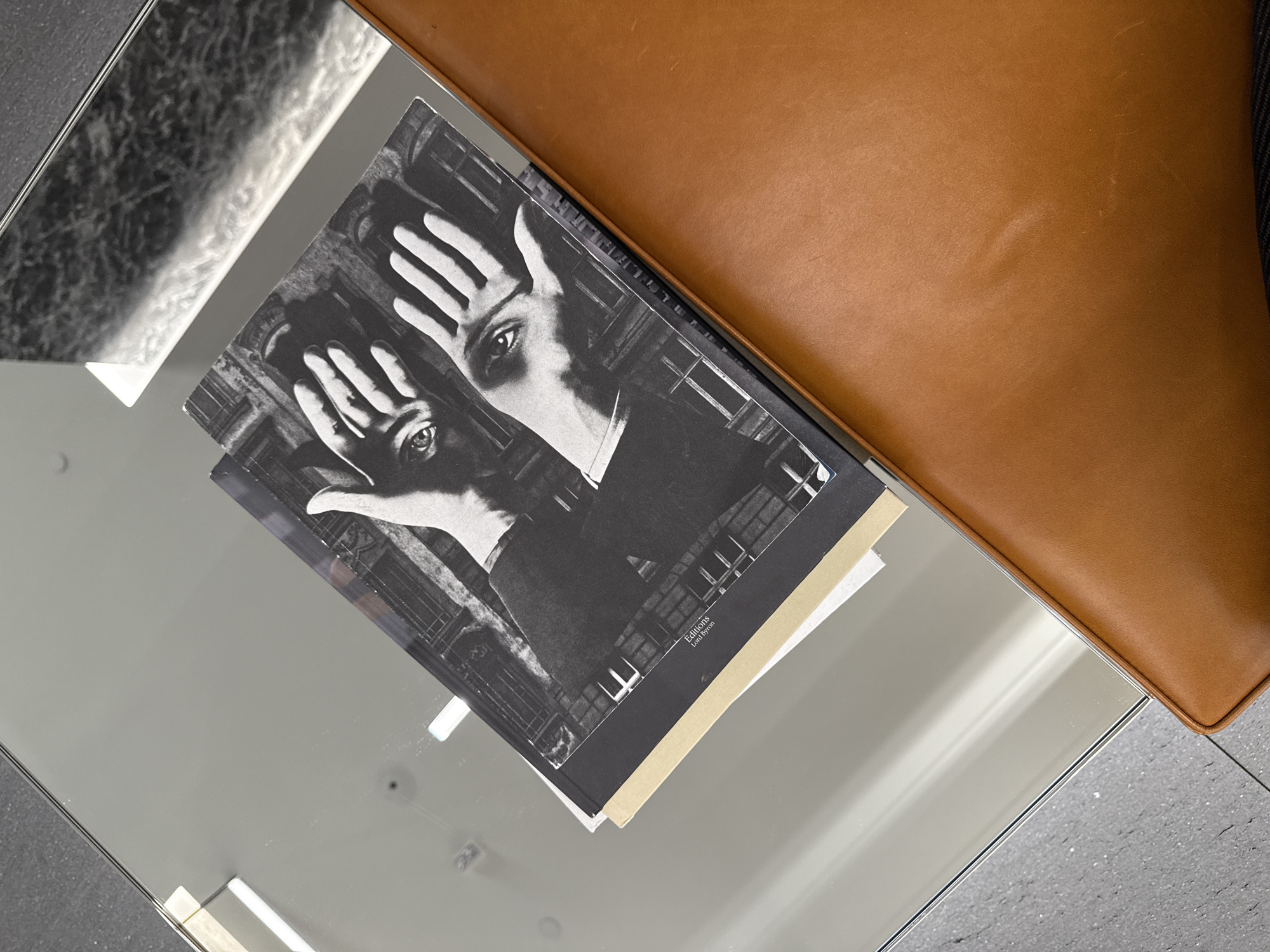 A high-angle, close-up shot depicts a glass table surface, upon which rests a book with a black-and-white cover image of a face with hands obscuring the eyes. Part of a brown leather cushion or chair is positioned on the right side of the frame. The setting appears to be indoors, likely within a domestic or commercial interior space in the city of San Diego, United States of America.