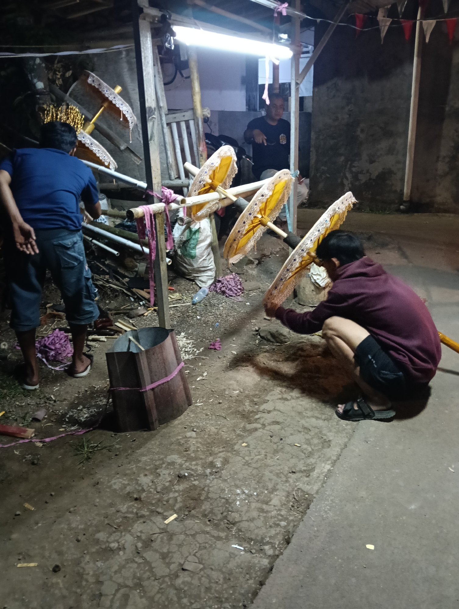 The image depicts an outdoor scene at night, possibly in a rural or semi-urban area of Tasikmalaya, Indonesia, characterized by a slightly uneven, dirt-and-gravel ground surface. In the foreground, a person wearing a dark maroon hoodie and black shorts crouches, holding a yellow umbrella with decorative white trim. To their left, another person in a blue t-shirt and denim shorts stands, facing away from the camera and towards several yellow umbrellas propped on a wooden structure.
Behind the standing person, a rustic wooden shelter with a corrugated metal roof is visible, illuminated by a bright overhead light. In the background, another individual, wearing a dark t-shirt, stands near the shelter, appearing to interact with something out of frame or engage in conversation.
The environment is cluttered with various objects, including bamboo poles, pink fabric tied to the wooden structure, a large burlap sack, and discarded wood scraps. A cylindrical wooden container with pink banding is positioned near the center of the frame. The overall impression is one of preparation or activity taking place in a functional, possibly workshop-like, outdoor setting at night.