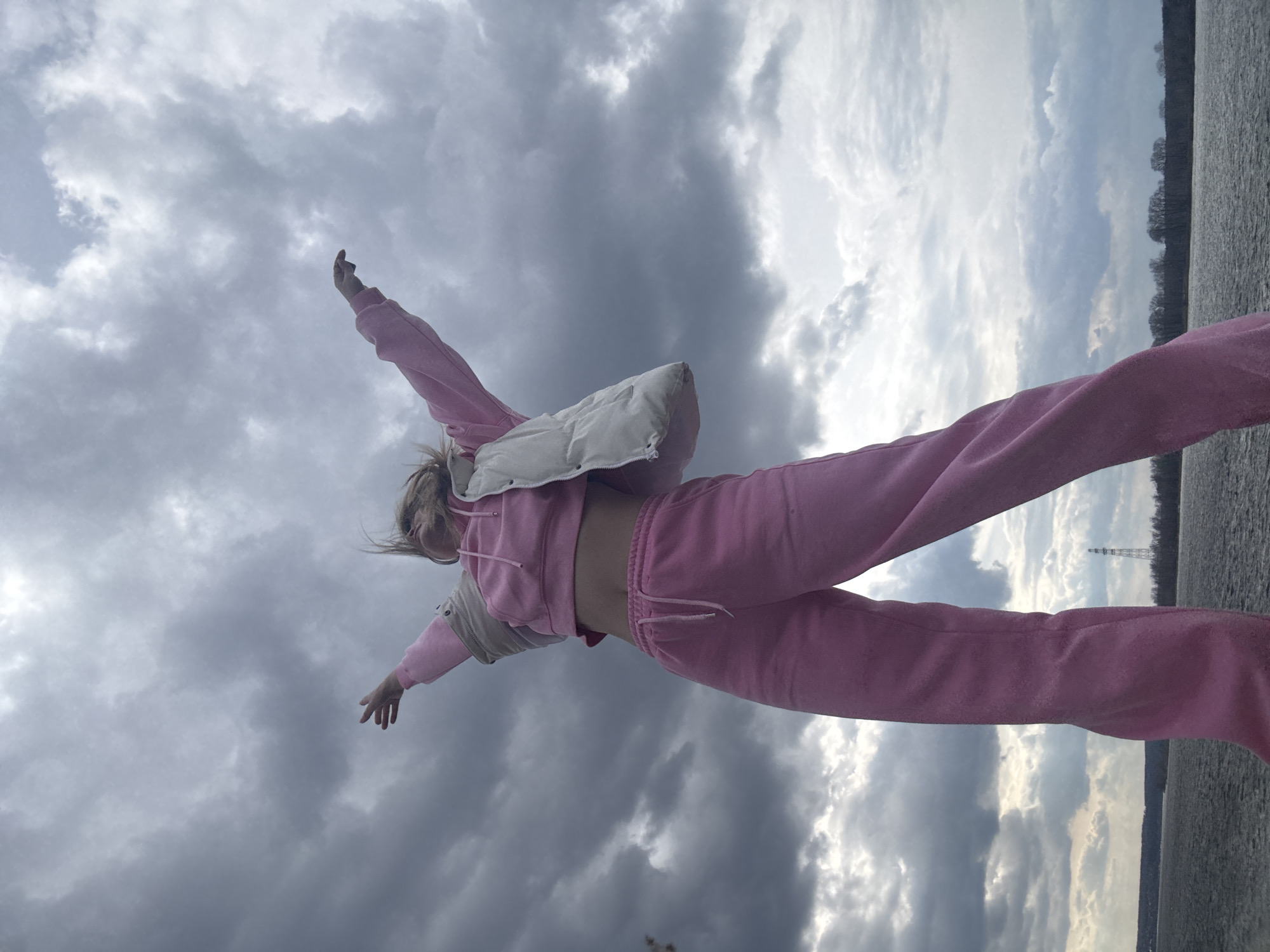 A low-angle outdoor shot captures a person posing with outstretched arms against a dramatic, overcast sky and a body of water. The individual, likely female, is positioned centrally, with their head tilted slightly back and blonde hair appearing wind-swept. They are dressed in a casual, two-piece pink tracksuit – a hooded top and sweatpants – worn over a light-colored, possibly off-white or light grey, puffer vest. A bare midriff is visible between the top and pants.
The background is dominated by a wide expanse of sky filled with heavy, dark grey clouds, interspersed with brighter patches where sunlight may be breaking through or reflecting. Below the horizon, a vast body of water, likely a lake or large river, stretches across the lower portion of the frame, its surface appearing textured. A distant treeline or landmass runs along the horizon. On the far right, a tall, slender metal structure, possibly a power transmission tower, rises above the distant trees.
The person's pose, with arms extended, conveys a sense of freedom, exhilaration, or an embrace of the expansive natural environment. The scene takes place under dynamic weather conditions, with significant cloud cover, likely in Chaikovsky, Russia, along the Kama River or a reservoir. There is no visible text in the image.