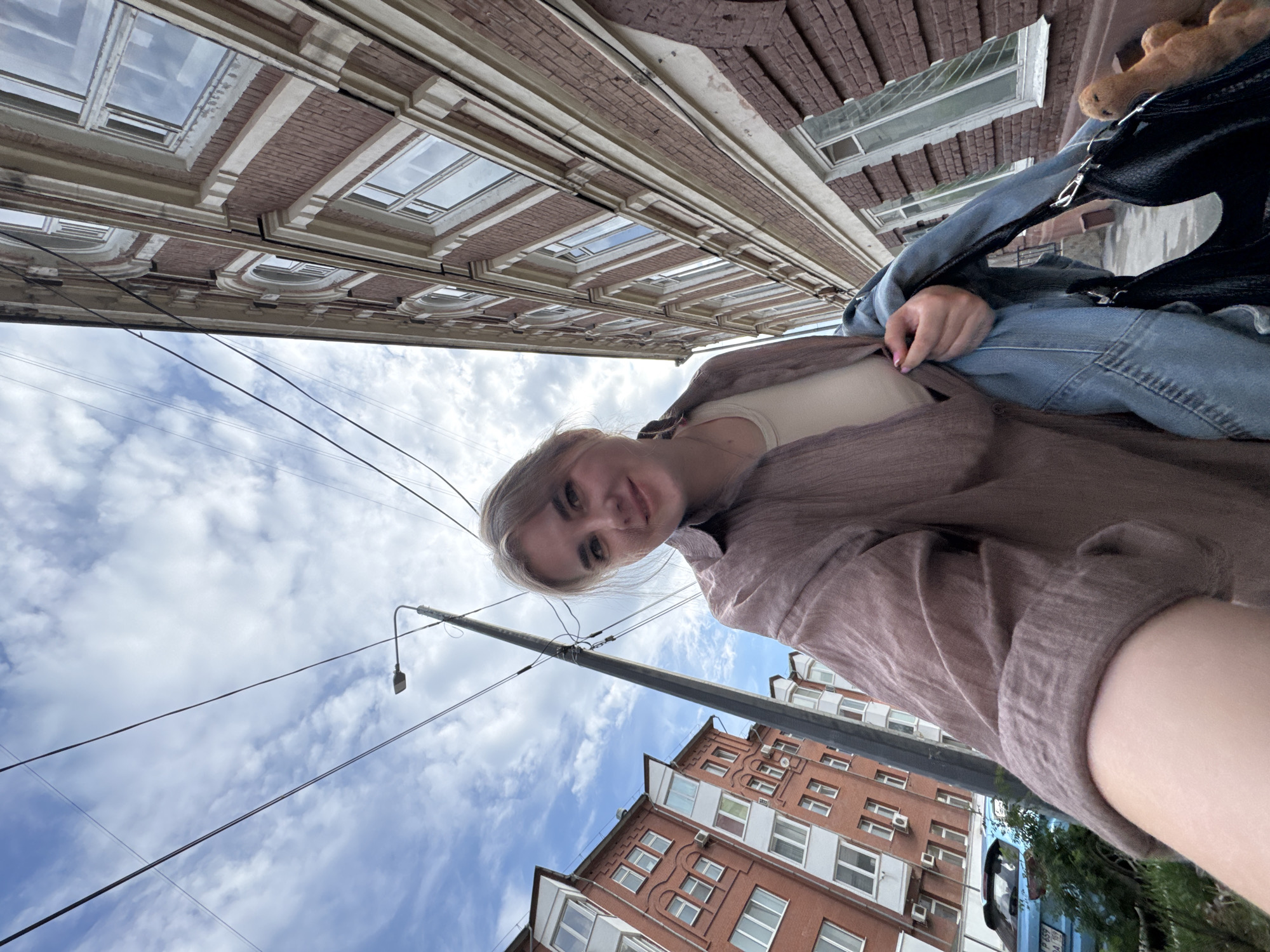 A low-angle, rotated selfie captures a young woman on a city street in Perm, Russia. She is in the foreground, looking directly at the camera with a soft smile. She has light-colored hair and eyes, and is wearing a light-colored top beneath a loose, textured brown or mauve shirt and blue jeans. Her left hand is visible near her midsection. A black shoulder bag with silver hardware is slung over her shoulder, from which a light brown plush toy, possibly a giraffe, peeks out.
The background features a bright, overcast sky with scattered white clouds. Power lines crisscross the upper part of the frame, and a lamppost with a street light fixture is visible to the right. Urban buildings line the street. To the left, an older building with ornate brickwork, decorative cornices, and rectangular windows reflects the sky. To the right, a multi-story red brick building with numerous windows and air conditioning units is visible. A portion of a blue vehicle, possibly a car or van, can be seen at the bottom right, with some indistinct white text on its side, likely a license plate, which is illegible.
The scene depicts a daytime urban environment under cloudy but bright weather conditions. The casual pose and gentle smile suggest a relaxed moment. The low camera angle creates a unique perspective, emphasizing the sky and the height of the surrounding buildings in Perm.
