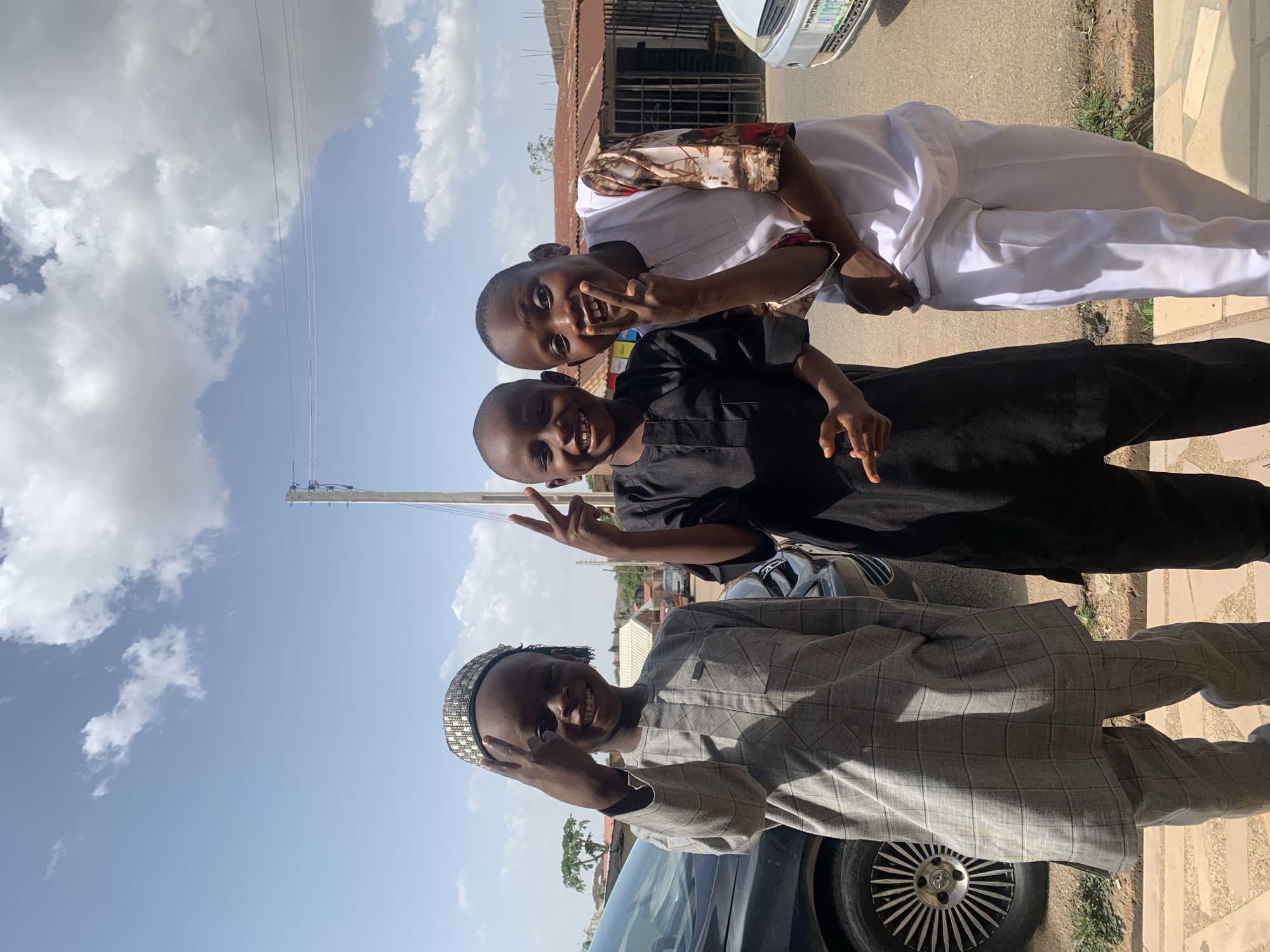 Three young boys in Jos, Nigeria, pose for a photo outdoors during the day. The sky is blue with white clouds. The boy in the foreground wears a gray checkered outfit and a beaded hat, standing in front of a dark car. The middle boy wears a black outfit, and the boy in the back wears a white outfit with a patterned top. All three are smiling and making peace signs with their hands. A utility pole stands in the background. Behind the boy in white is a building with a rusted metal roof. A silver car is parked on the street.