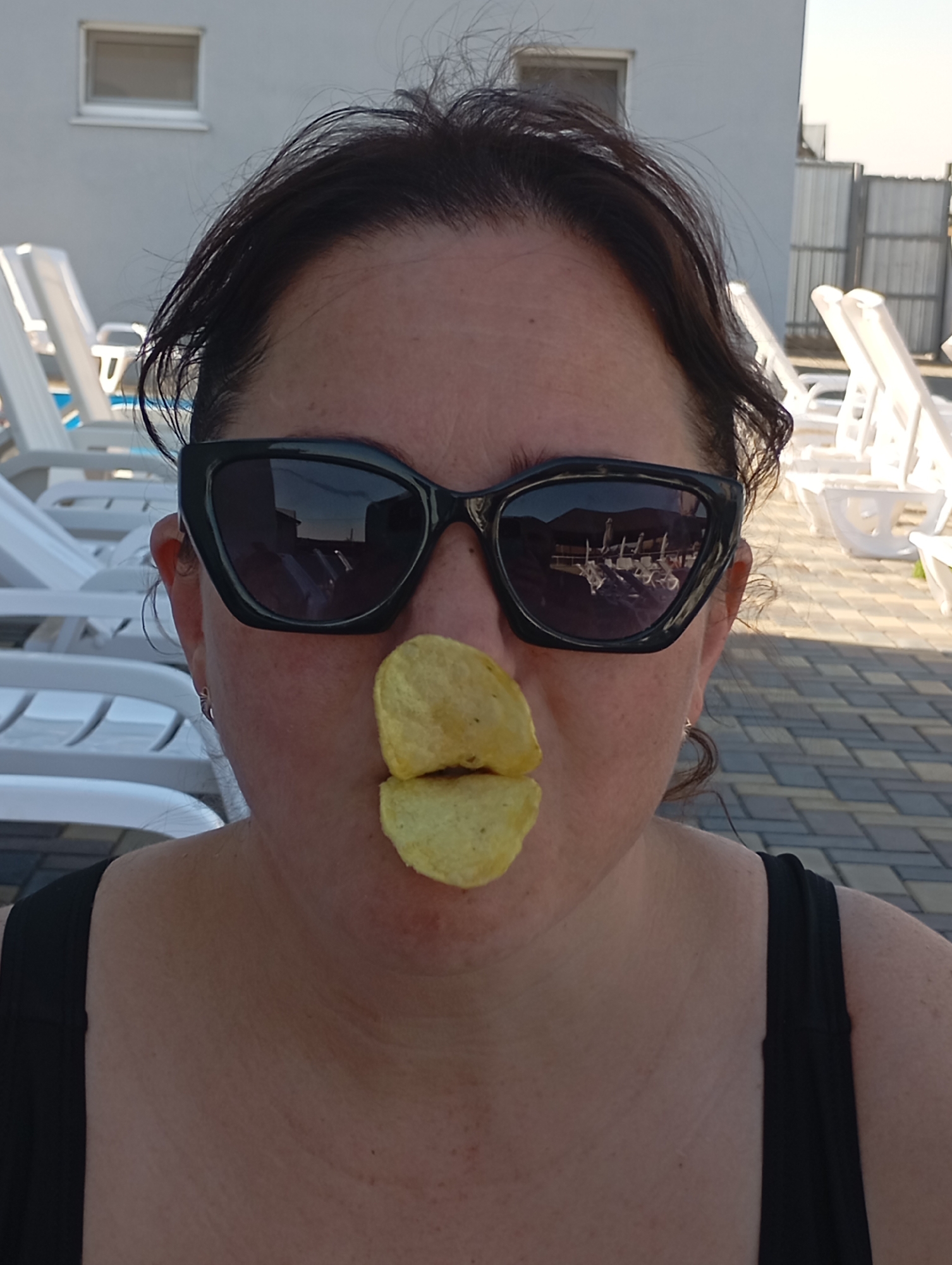The media file shows a close-up shot of a woman in an outdoor setting, possibly a resort or pool area, given the presence of lounge chairs and paved ground. The woman is wearing large, black sunglasses, which reflect a scene with more lounge chairs and a building. Two potato chips are placed on her lips, giving the appearance of a playful or humorous expression. Her hair is dark and styled loosely. The background suggests a sunny day, and the overall mood appears lighthearted and casual. The woman is wearing a black tank top. The location is identified as Vladimirovka, Moldova. No visible text is present in the image.