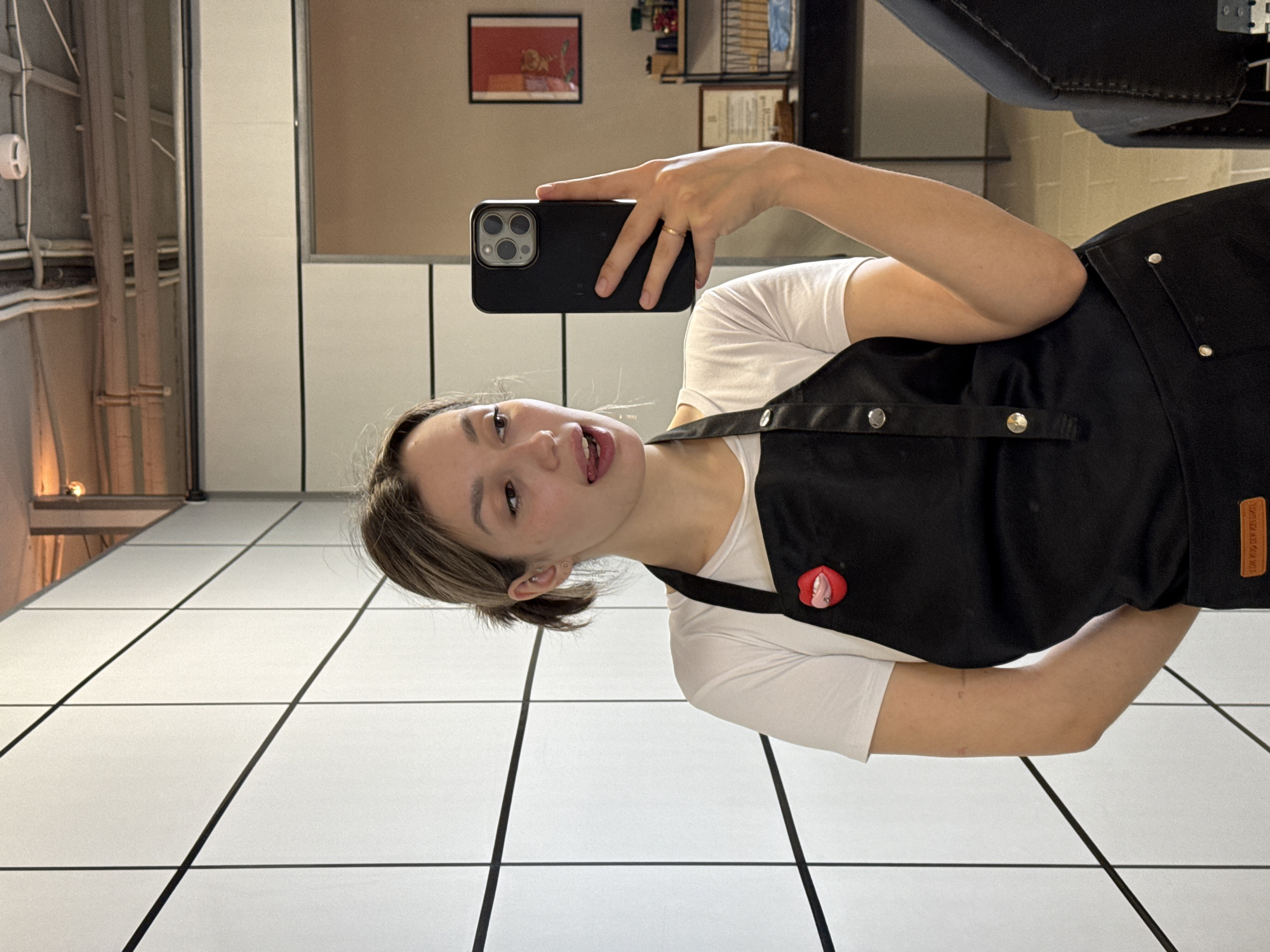 A young woman is captured in a rotated selfie, holding a dark-cased smartphone with a visible triple-lens camera array in her right hand, pointing it towards herself. She wears a white short-sleeved t-shirt and a black apron adorned with silver buttons or rivets. A distinctive red and pink lip-shaped pin is affixed to her apron. Her light brown hair is pulled back, and she has a ring on her right ring finger. Her expression is playful, with her mouth slightly open and her tongue visible.
The setting appears to be indoors, possibly a hair salon or a similar commercial establishment. The floor is covered in a prominent white tile pattern with black grid lines. The background features light-colored walls, a framed red artwork, and what looks like industrial-style exposed pipes and possibly ventilation on the ceiling. A large, dark piece of furniture, potentially part of a salon chair or station, is visible in the upper right. On a brown tag attached to her apron, the text "LIVE LIFE YOU DESERVE" is partially visible. The overall lighting suggests an indoor daytime setting.