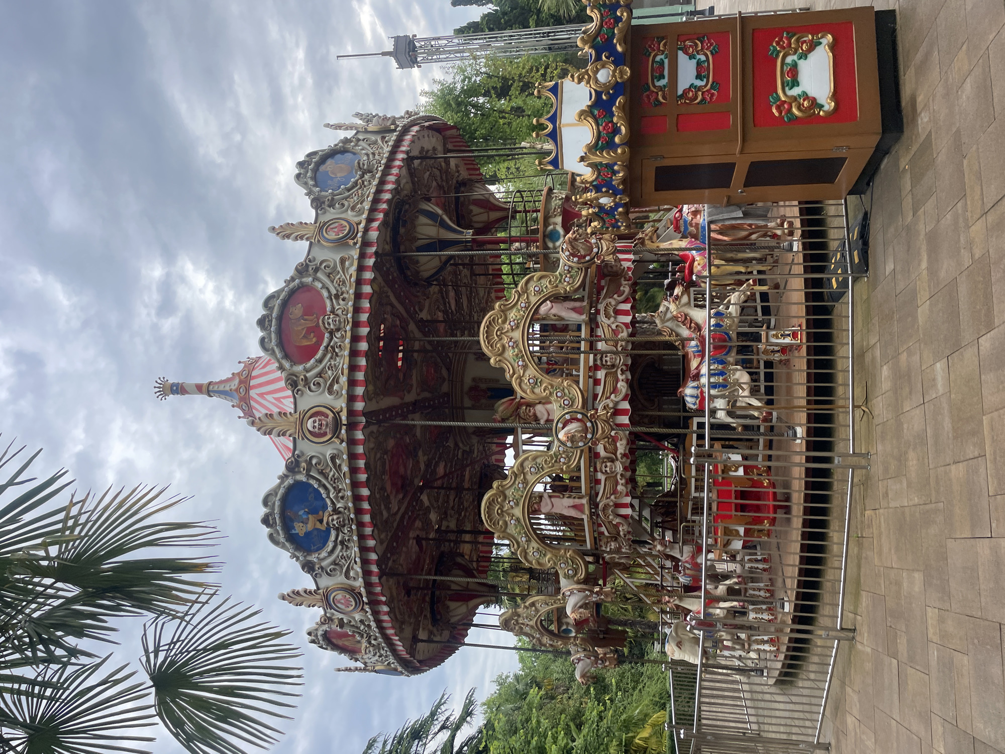 The image depicts a carousel in Sochi, Russia. It is a highly decorated carousel with numerous horses and ornate carvings. The carousel is fenced in by a silver metal fence. The ground around the carousel is paved with large, square stones. In the background, there are trees and a tall tower. The sky is overcast. In the foreground, palm leaves can be seen. The image is rotated by 90 degrees.
