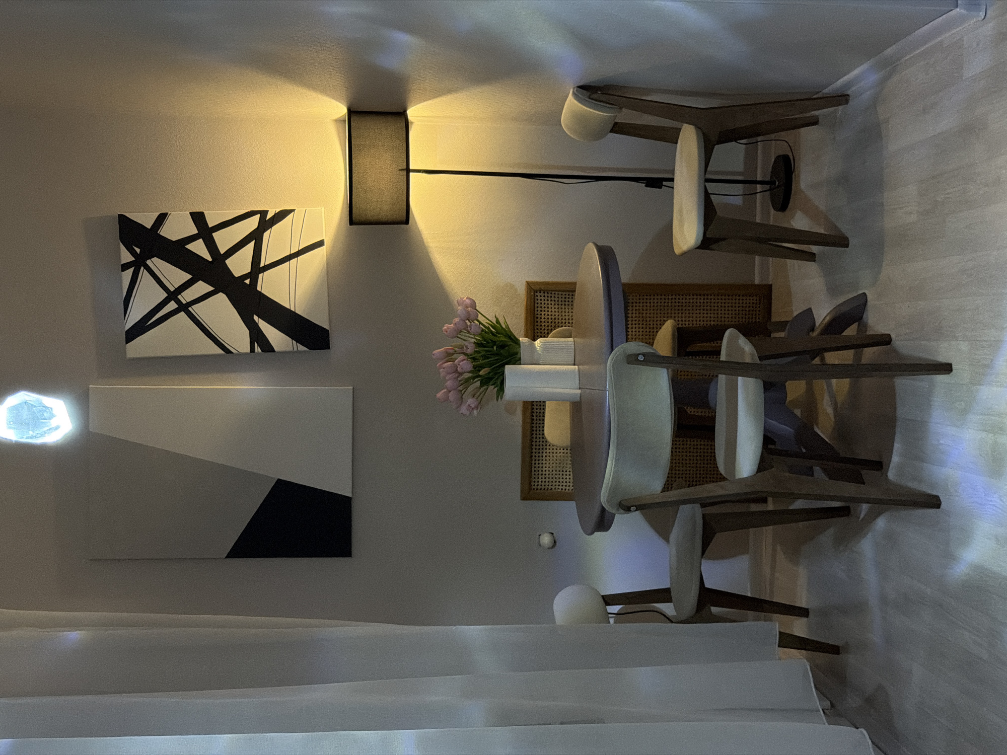 The image depicts a cozy dining nook within an apartment, likely located in Moscow, Russia. The scene is set in the evening, as indicated by the warm glow of a floor lamp illuminating the wall and casting shadows.
Central to the image is a small, round dining table with a muted purple top, surrounded by four wooden chairs with light-colored upholstered seats and backs. One chair is positioned in the foreground, while three others are partially visible around the table. A woven rattan chair back is also visible behind the table.
On the table, a white vase holds a bouquet of pink tulips, adding a touch of color and freshness to the otherwise neutral palette of the room. The walls are a light, neutral gray, adorned with two pieces of abstract art. The left artwork features geometric shapes in black, white, and gray, while the right piece consists of bold black lines on a white background.
To the left, a sheer white curtain drapes, partially obscuring a white globe lamp on a stool. The flooring is a light-colored wood laminate, with subtle reflections of light from the lamp and an unseen light source near the ceiling, possibly a ceiling fixture or a window with interesting light effects. The overall impression is one of a comfortable and aesthetically pleasing interior space, designed for intimate gatherings or quiet meals. There are no people visible in the image.