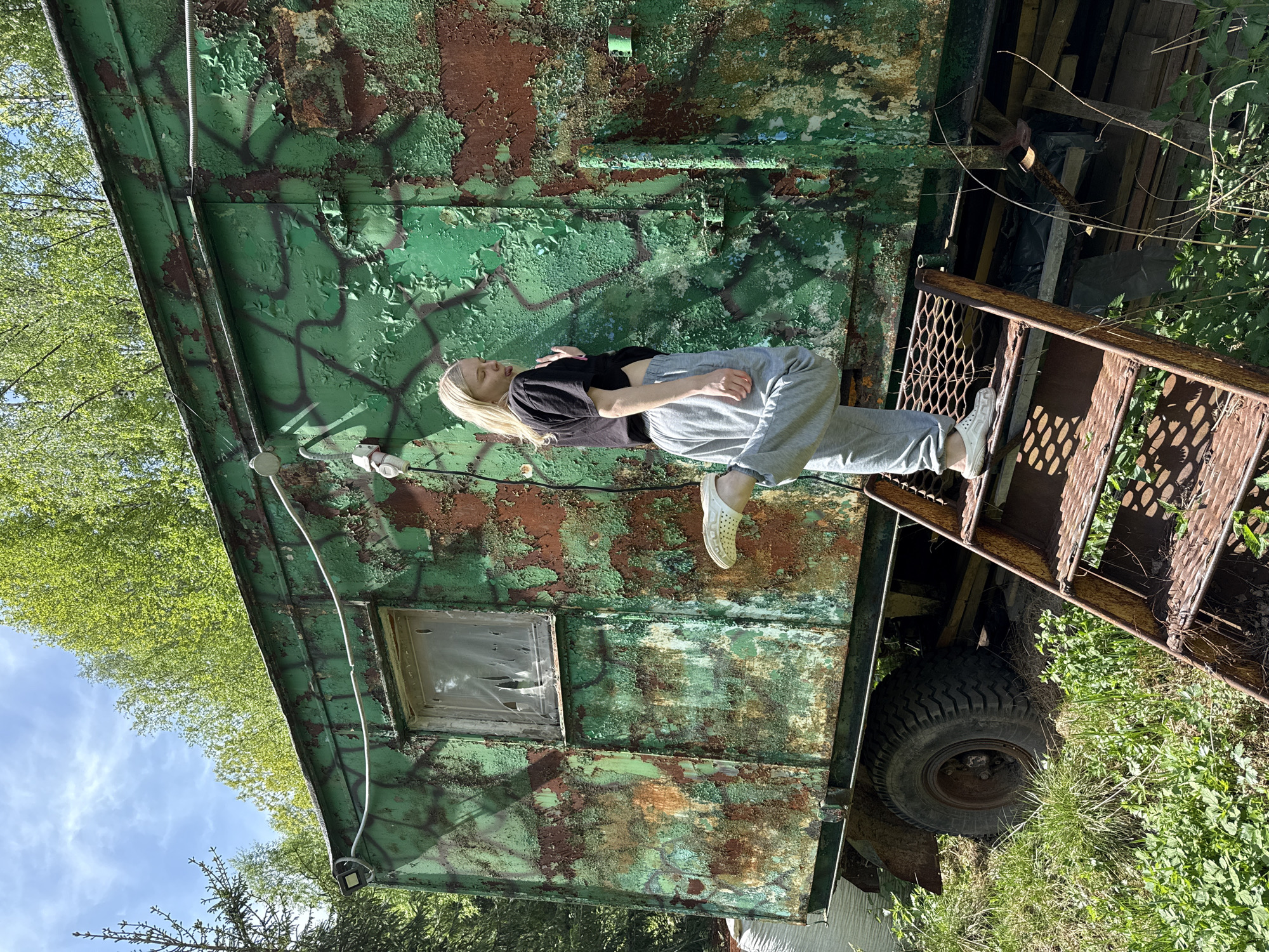 A young woman with long blonde hair, wearing a black crop top and gray sweatpants with white shoes, is posing on the rusty stairs attached to an old green and brown camouflage-painted truck in what appears to be a wooded area. The truck has a window boarded up and is surrounded by green grass and trees. The photo was taken on a sunny day. There is no indication that this scene is in Zhigalki, Russia.