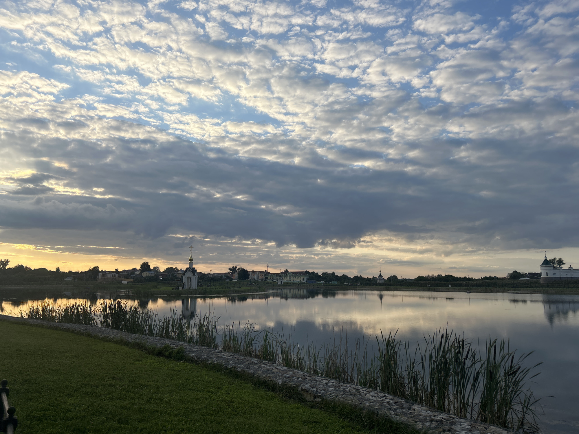 This image captures a serene waterscape in Diveyevo, Russia, likely during the golden hour. The foreground features a well-maintained grassy lawn bordered by a low stone wall. Reeds and cattails grow along the edge of the tranquil body of water that dominates the midground. The water's surface acts as a mirror, reflecting the sky and the structures on the opposite bank.
On the far side of the water, a line of buildings and religious structures is visible. A prominent white church with a golden dome and cross stands out on the left, its reflection clearly visible in the water. Further to the right, other buildings, including what appears to be a larger white structure with a distinctive turret, are also discernible. The skyline is framed by a dramatic sky filled with a mix of gray and white clouds, with patches of soft yellow and orange light suggesting the presence of the setting or rising sun. The overall atmosphere is peaceful and picturesque, indicative of a landscape often associated with holy sites.