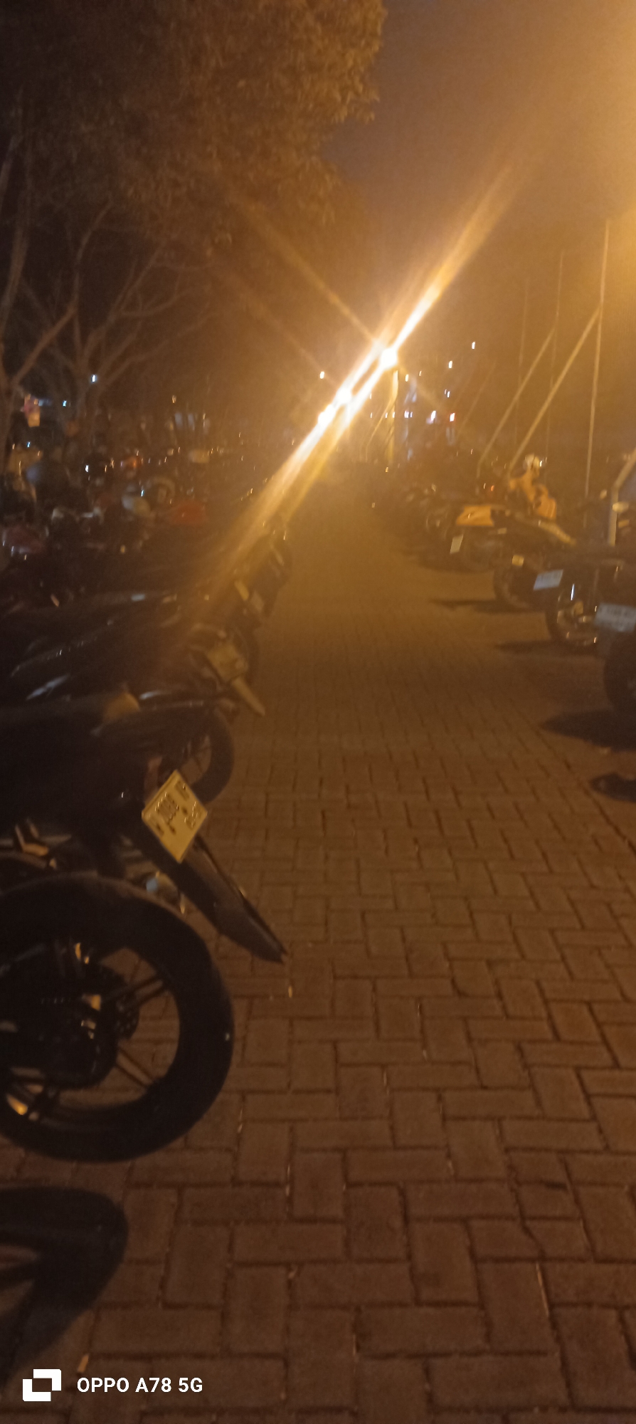 A nighttime outdoor scene shows a line of parked motorcycles and ATVs on a brick-paved surface, likely a parking lot. The area is illuminated by a bright overhead light source, creating strong glare and shadows. Trees line the left side of the image, their branches visible against the night sky. The ground is paved with rectangular bricks laid in a herringbone pattern. The motorcycles and ATVs are parked closely together, with their headlights and various components visible. Some of the vehicles have license plates. A building or structure with poles and cables is visible in the background, suggesting a public space. The overall atmosphere is dim and slightly hazy due to the artificial lighting. The location is identified as Pranti, Indonesia.