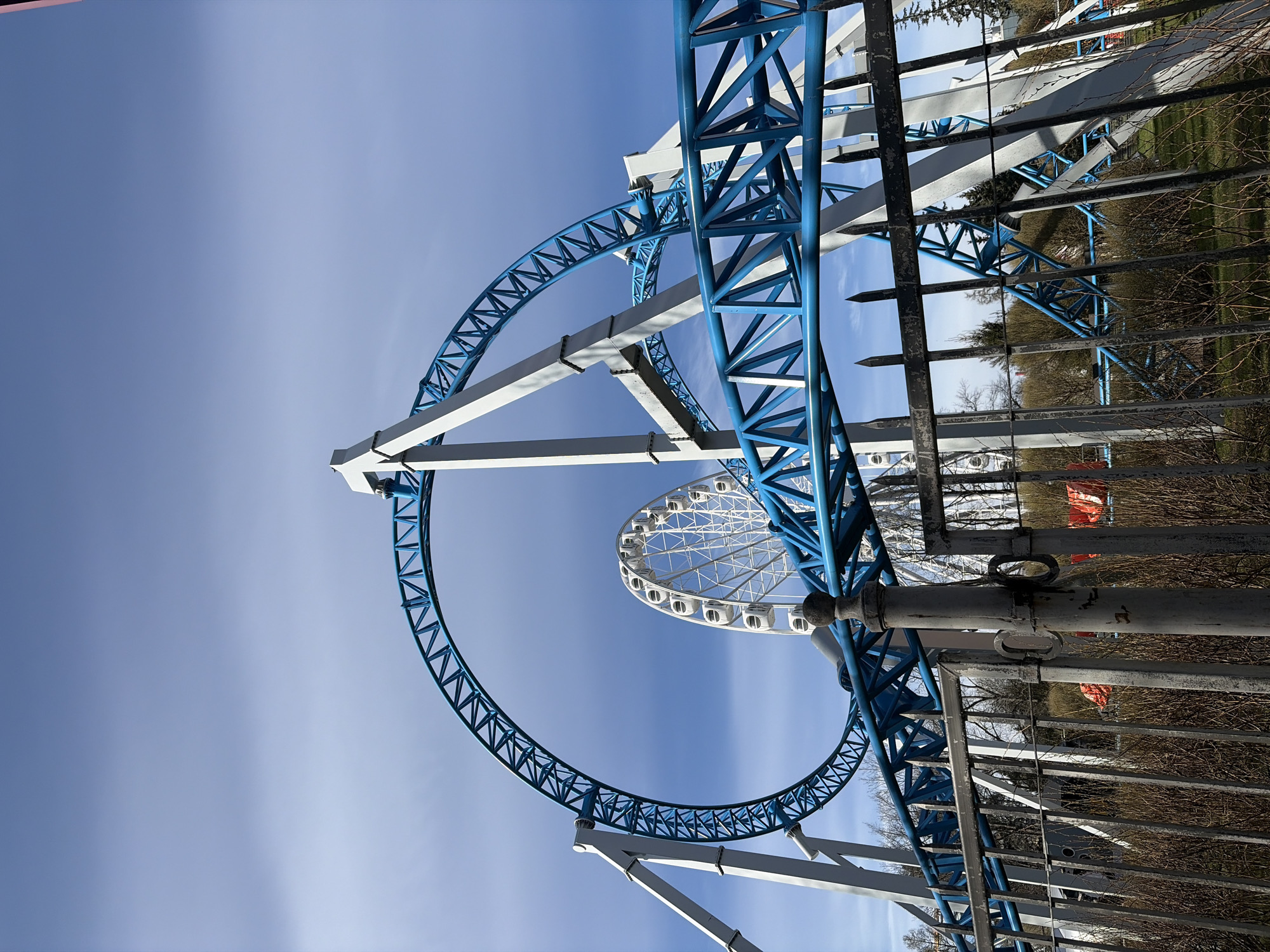 The image shows an upward view of a blue and white roller coaster with a Ferris wheel in the background. The sky is blue with light clouds. The roller coaster has a loop structure and is made of a blue metal framework with white supports. The Ferris wheel is white with enclosed gondolas. In the foreground, there is a rusty metal fence. The location is Saint Petersburg, Russia, identifiable from the presence of the roller coaster and the Ferris wheel, which are part of a local amusement park. There are no people visible in the image. The overall environment appears to be a clear, sunny day.