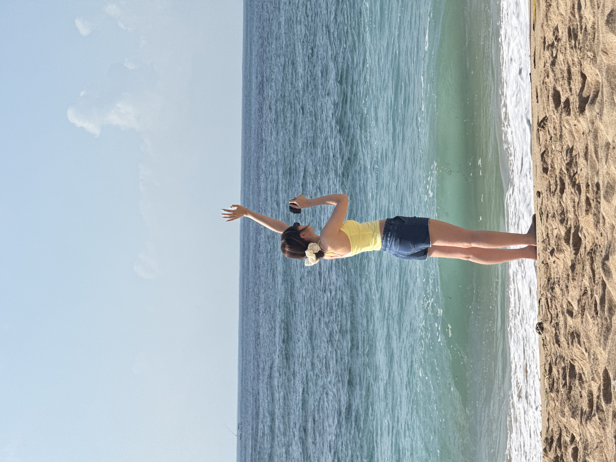 A person stands on a sandy beach in Kuta Selatan, Indonesia, facing the ocean. The person is wearing a yellow tank top and denim shorts, with a large white scrunchie in their hair. They are holding a black camera in their right hand and have their left arm raised, with their hand open. The ocean is a deep blue with turquoise waves crashing on the shore. The sky is a pale blue with a few white clouds. The beach is made of light brown sand, with some debris scattered on the surface. In the distance, a thin, dark line suggests a boat or a distant landmass. The scene is bathed in daylight.