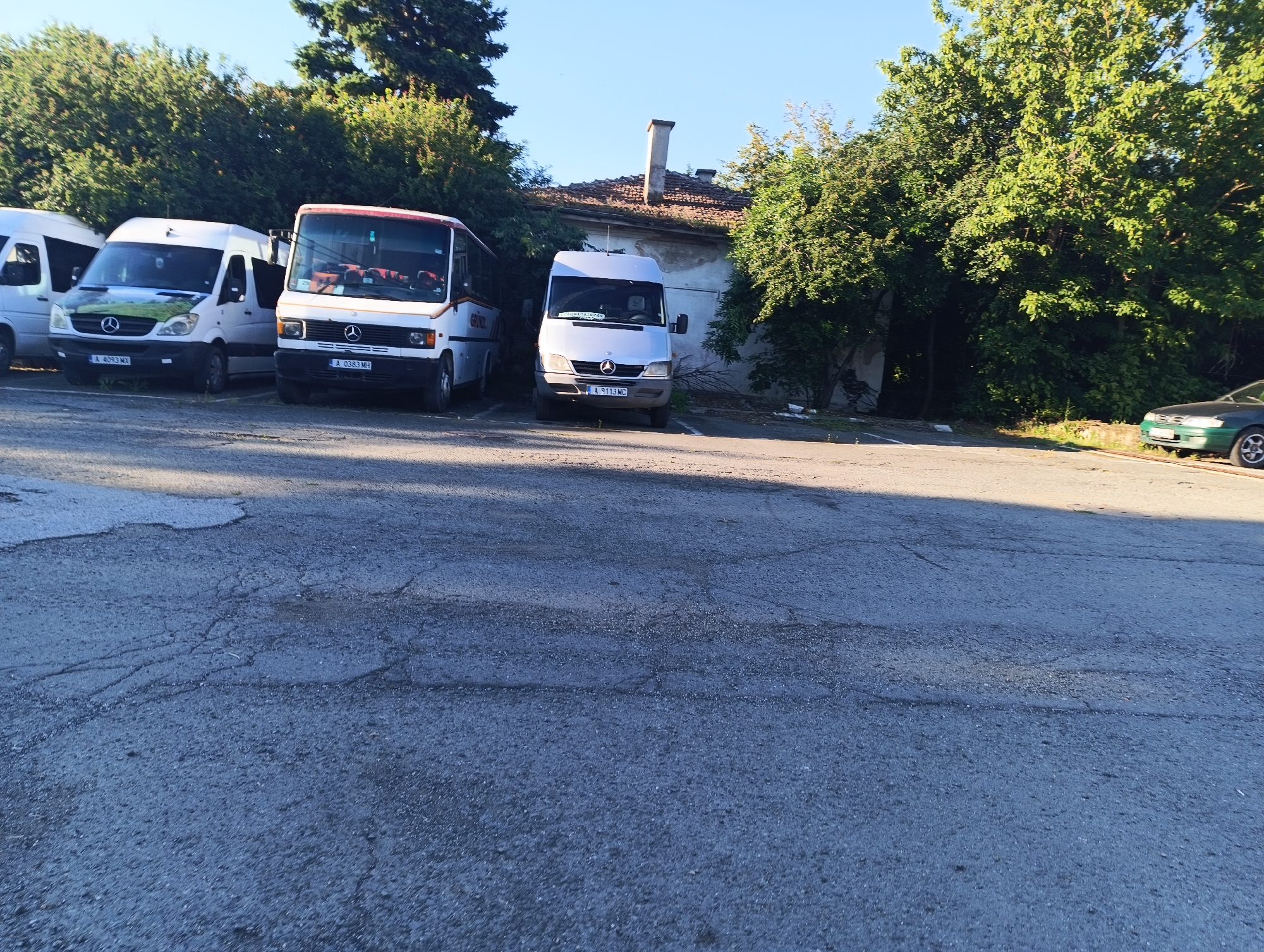 The image depicts a row of parked vehicles, including several white vans and a bus, on a cracked asphalt surface. The vehicles are situated in front of a building with a tiled roof and a prominent chimney. Dense green foliage surrounds the area, with tall trees to the left and right of the building, and more trees visible in the background. A dark green sedan is parked on the far right of the image. The scene appears to be a parking lot in an urban environment, possibly in Karnobat, Bulgaria, given the license plates on some of the vehicles. The lighting suggests it is daytime. No people are visible in the image.